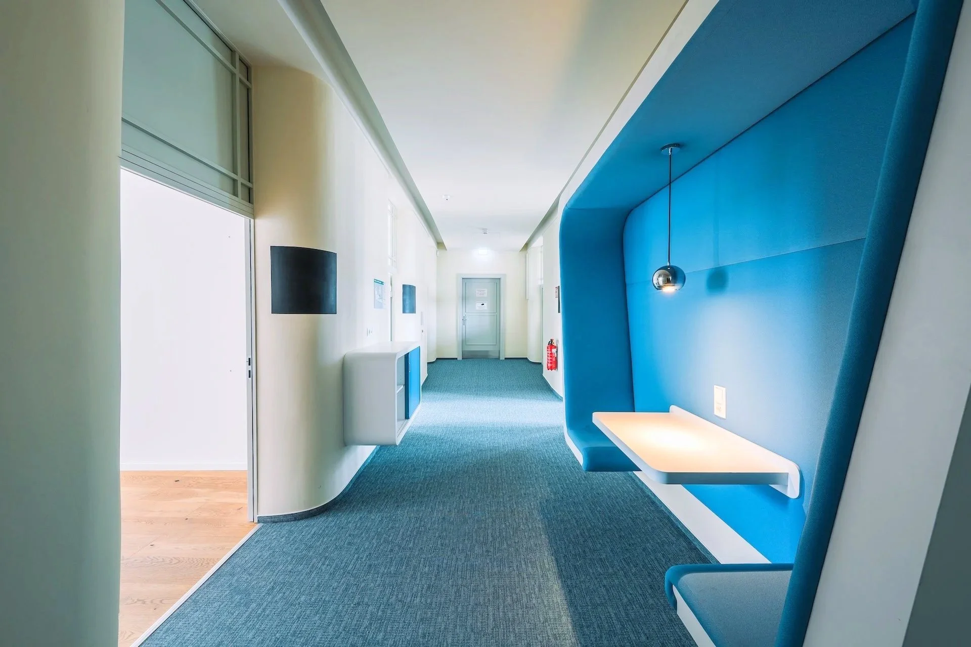 Modern corridor with cream walls, blue carpet, and a blue accent wall with a built-in white desk and spherical hanging light. There is a fire extinguisher and wall-mounted screens or art pieces along the corridor.