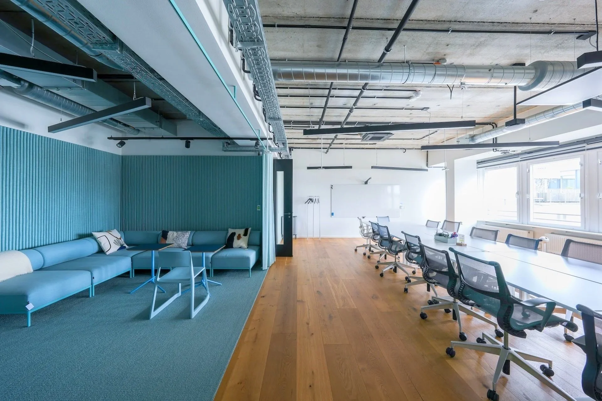 Modern coworking space with a seating area on the left with a blue sofa and pillows, and a long table on the right with multiple office chairs, large windows letting in natural light, and an exposed ceiling with ductwork and pipes.