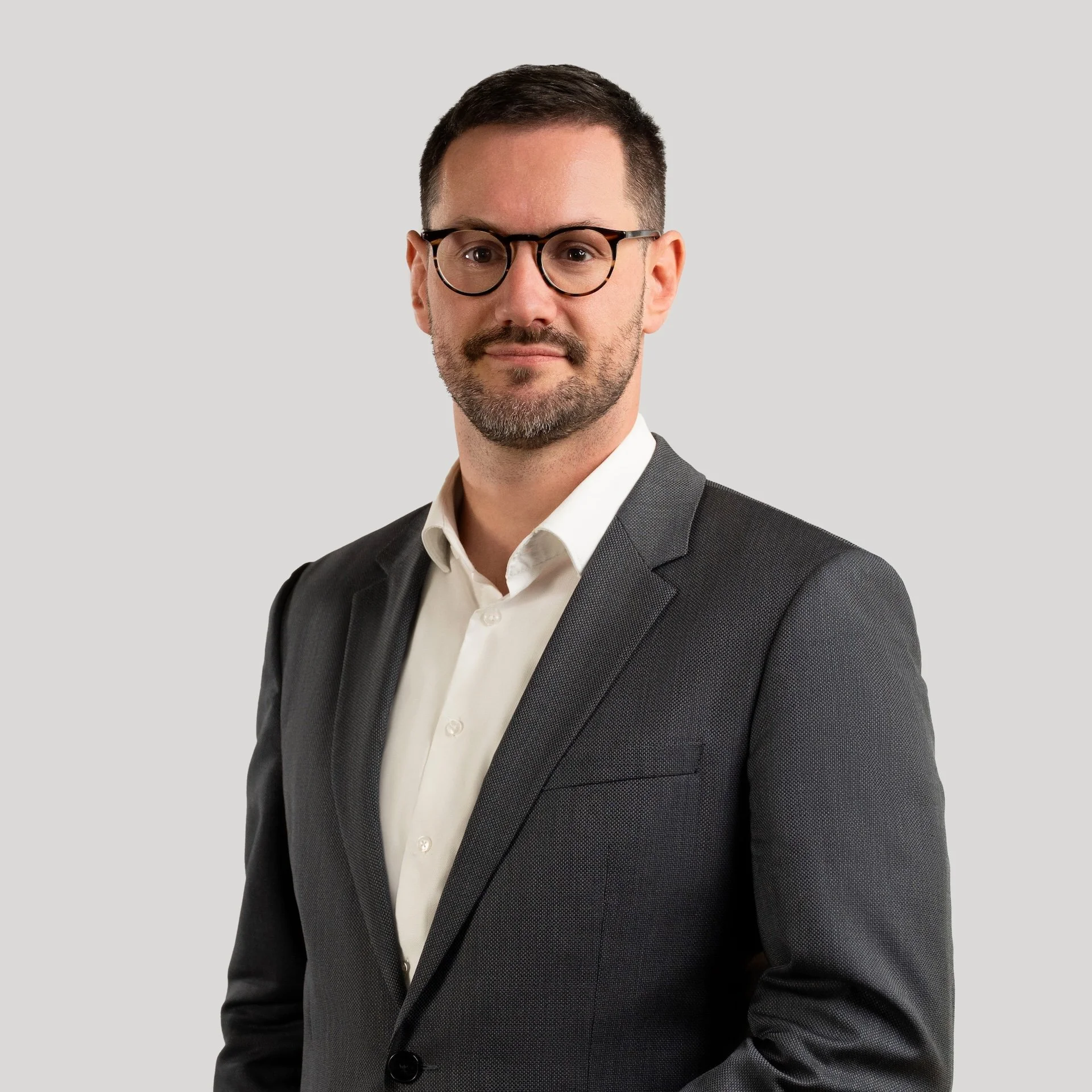 Portrait of a man with dark hair, glasses, a beard, wearing a dark gray blazer and white dress shirt against a plain light background.