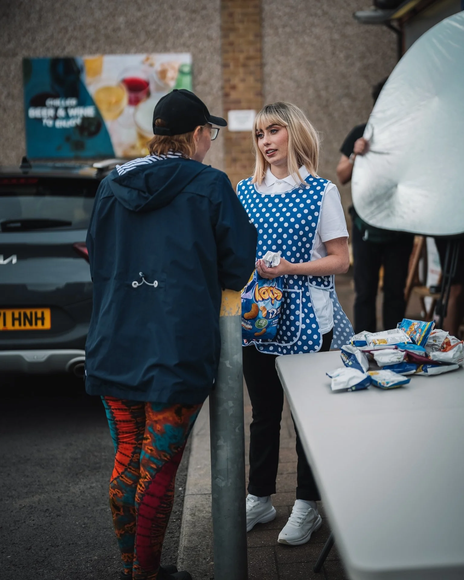 A young woman with blonde hair, wearing a blue polka dot apron over a white shirt, is talking to a person dressed in dark clothing and a cap, at an outdoor event. They are near a table with snack bags, with a car and a large advertising banner in the