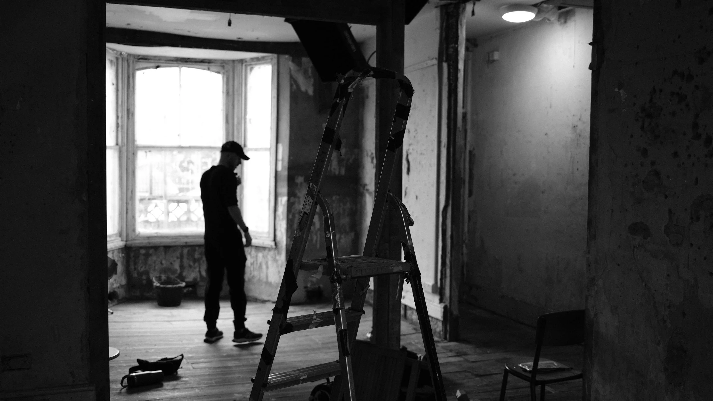 A person standing in a partially renovated room, looking down, with a ladder in the foreground and a large window in the background.