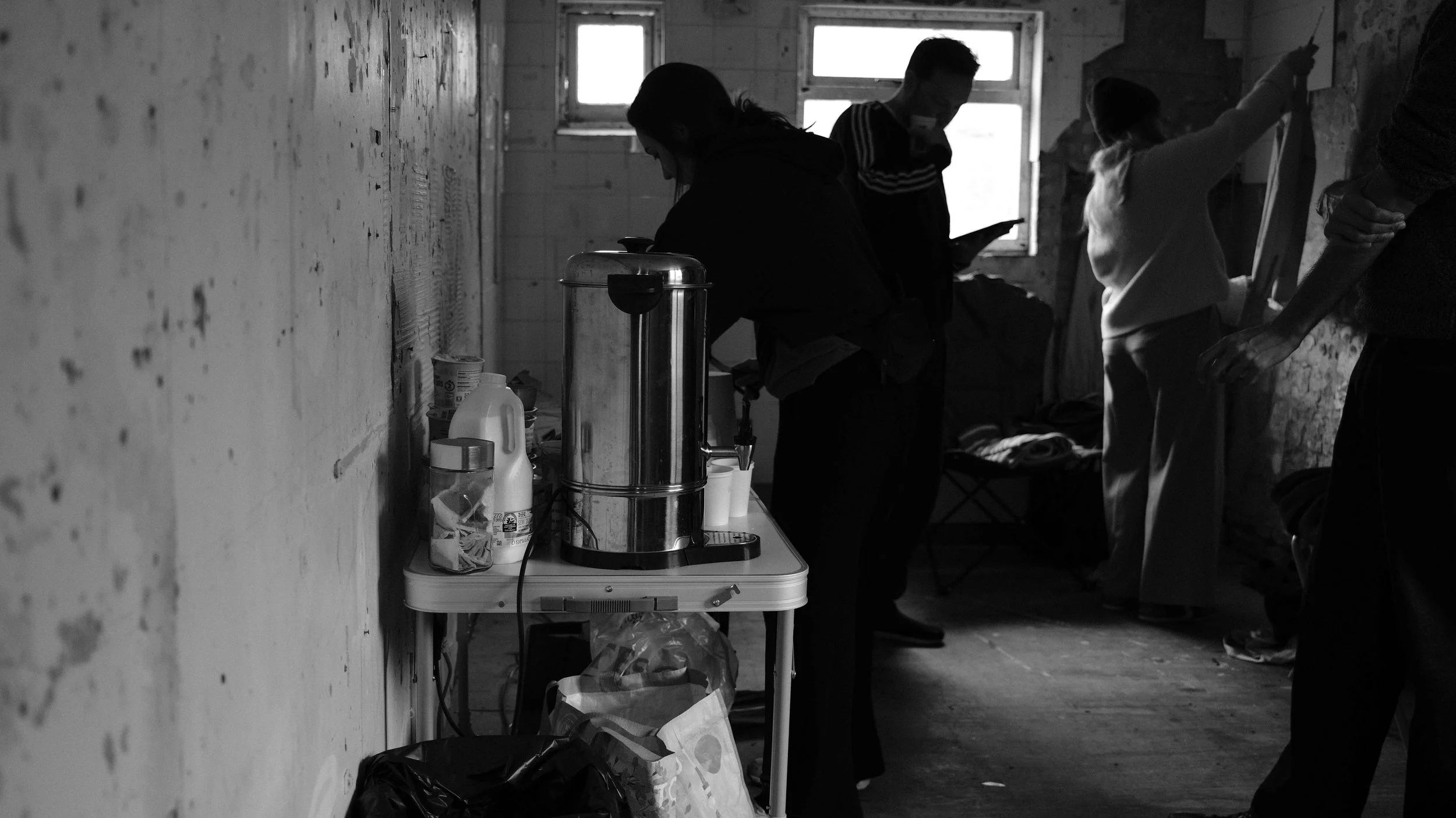 People in a dimly lit room with some on the left side near a table with a coffee dispenser, and others on the right side, with some writing or working on a wall. The room has old, worn walls and a wooden floor.