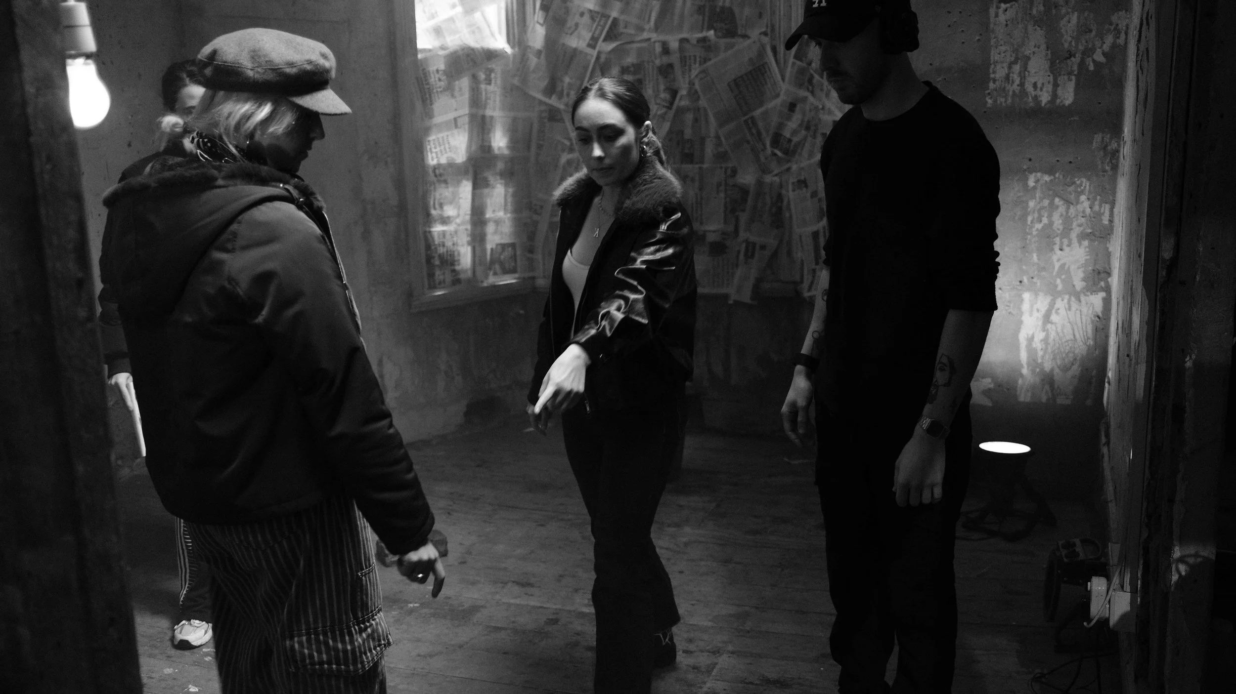 Three people standing in a room with rough concrete walls and newspapers taped to the window. One woman is pointing at something, another woman with a hat looks down, and a man stands with his hands by his side. A small light on the floor illuminates