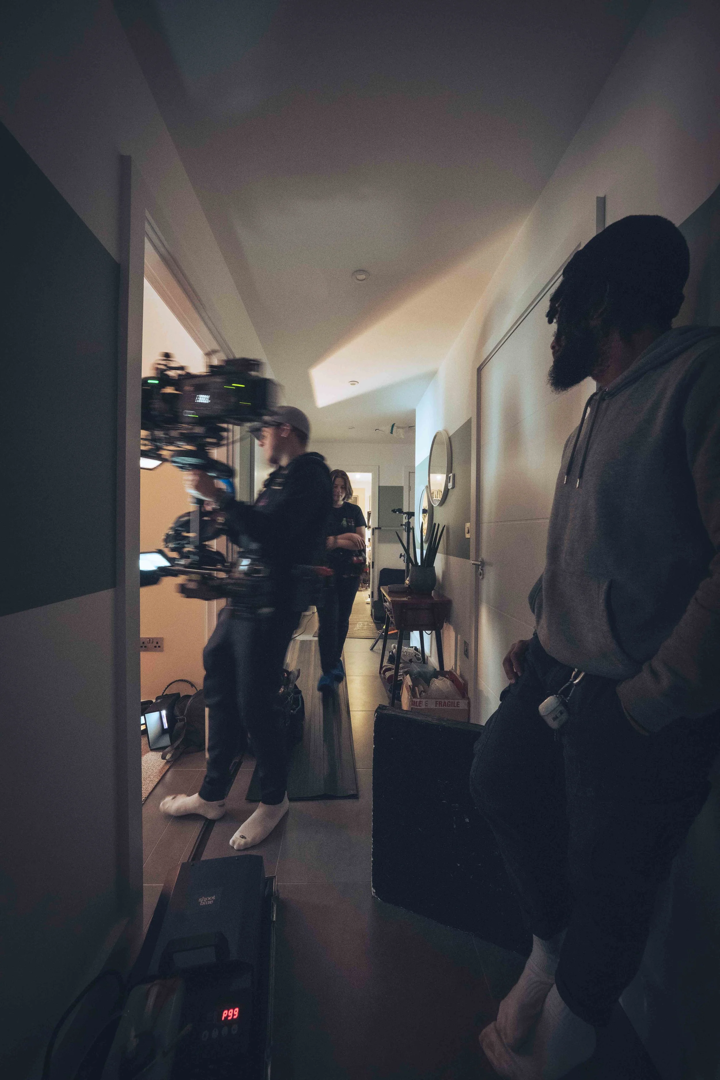 A group of people in a hallway, with one person holding a professional camera and tripod, while others watch or stand nearby. The scene appears to be part of a filming or photography setup.