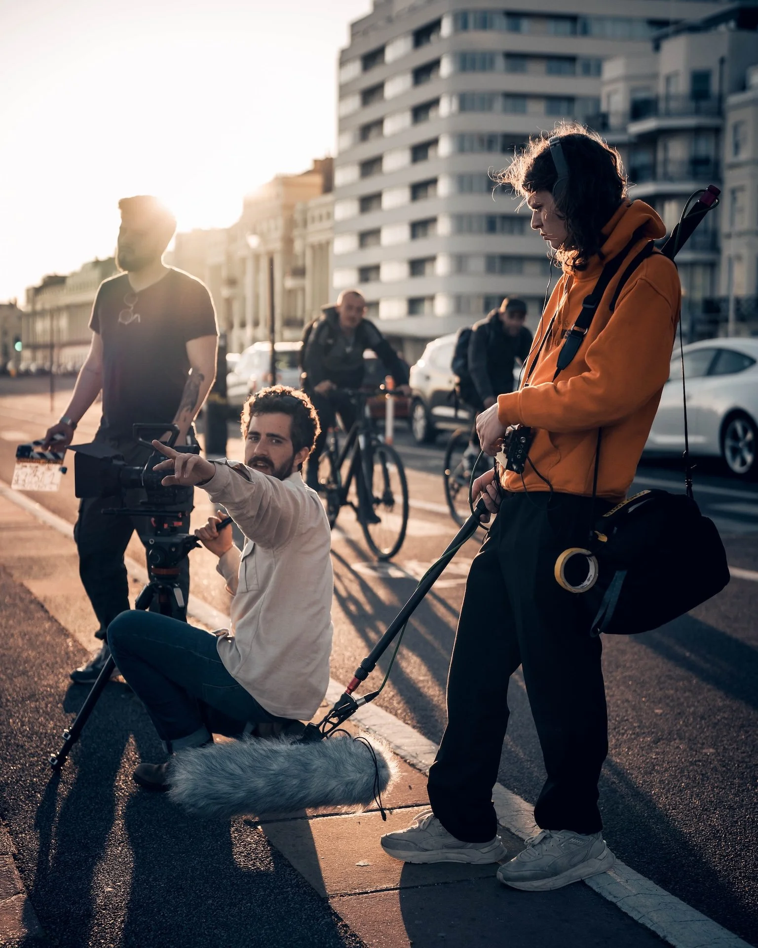 Two people on a city street at sunset, one in an orange hoodie with headphones and a backpack, the other crouched with a camera, and two more people riding bikes in the background.