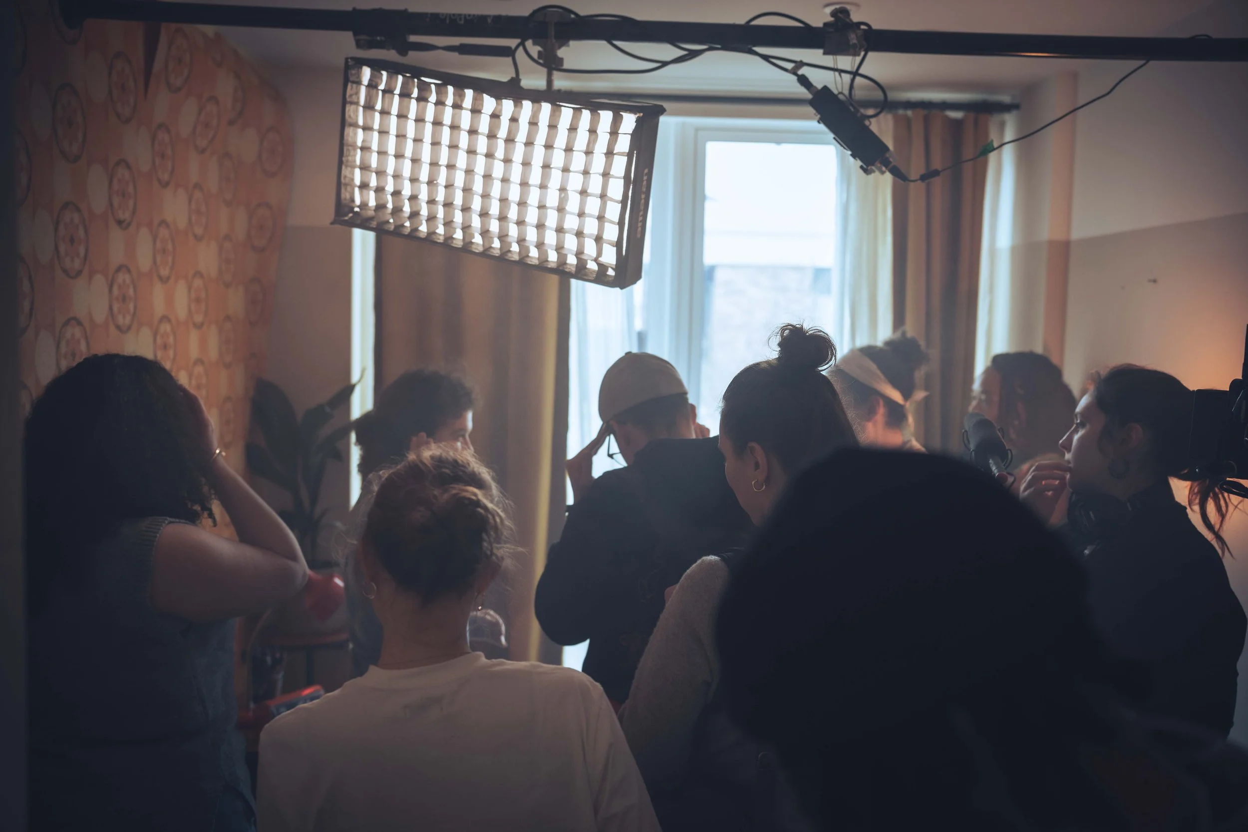 A group of people gathered indoors, with a large softbox light above, near a window with yellow curtains, and patterned wallpaper on the wall.