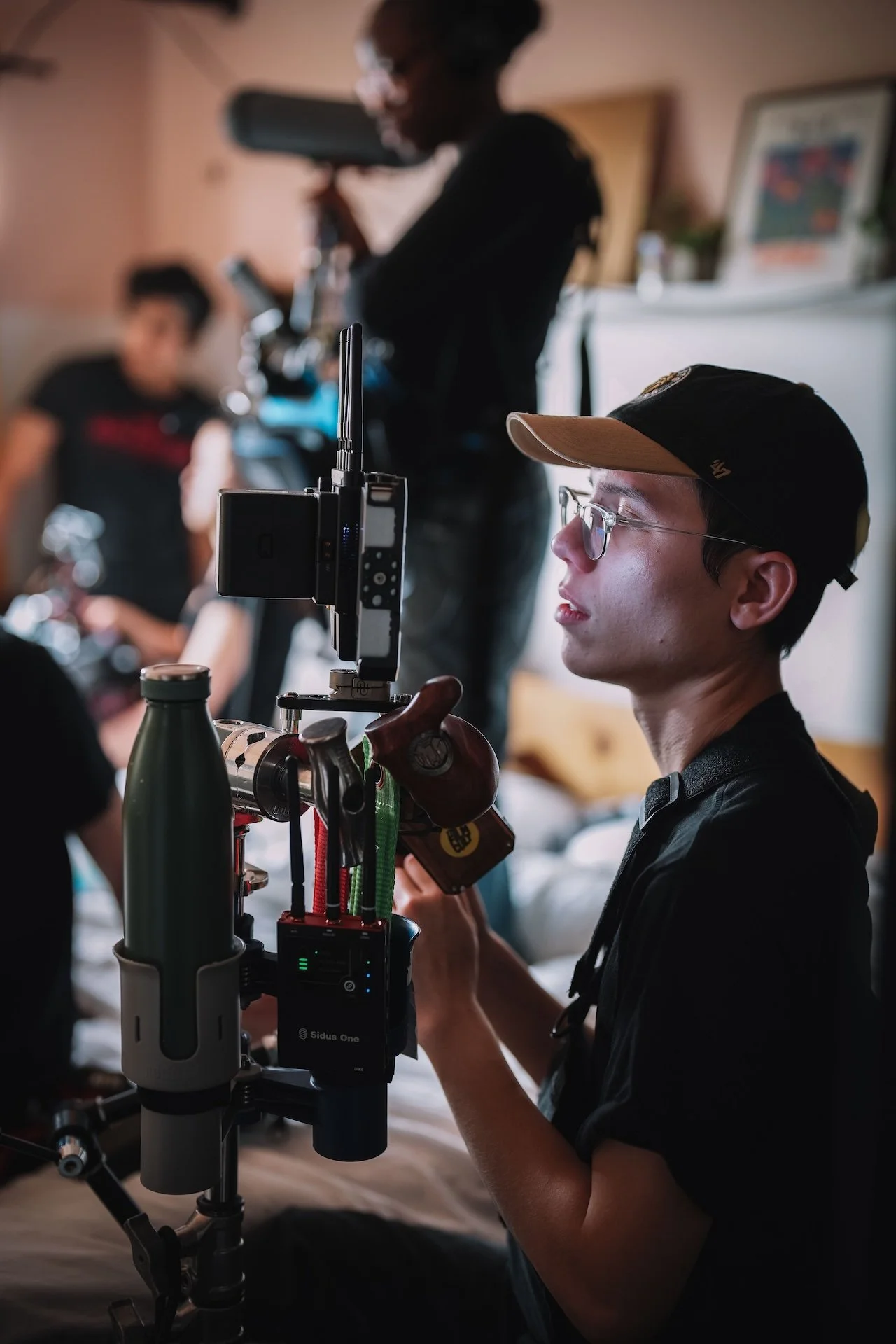 Young man with glasses and a cap operating a camera or filming equipment in indoor setting, with other people in the background.