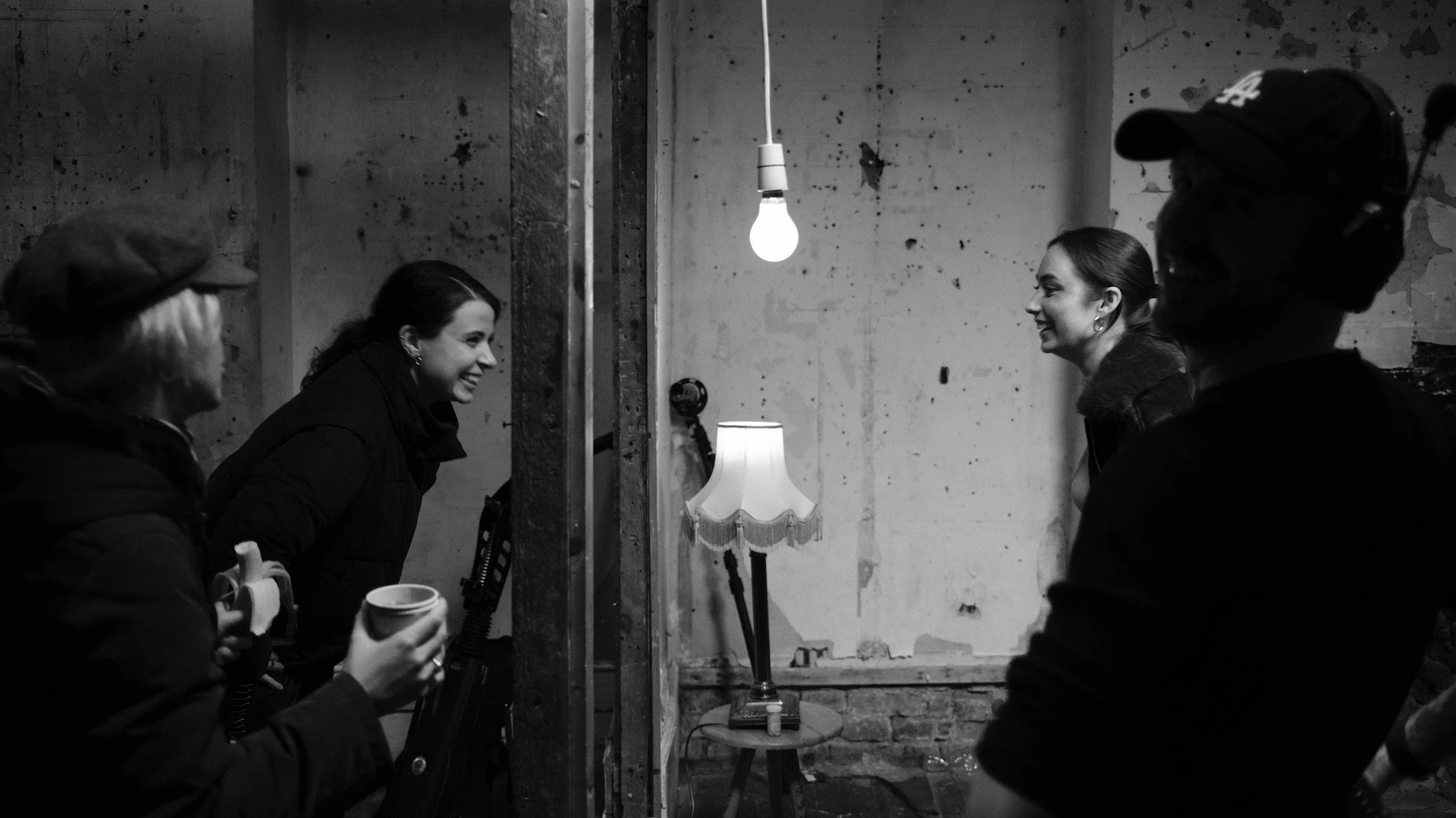 Four women are standing and smiling in a partially finished room with bare walls. Two of them are on the left side, holding mugs, and two on the right side, one of whom is wearing headphones and a cap. There is a lamp with a fringed shade on a small 
