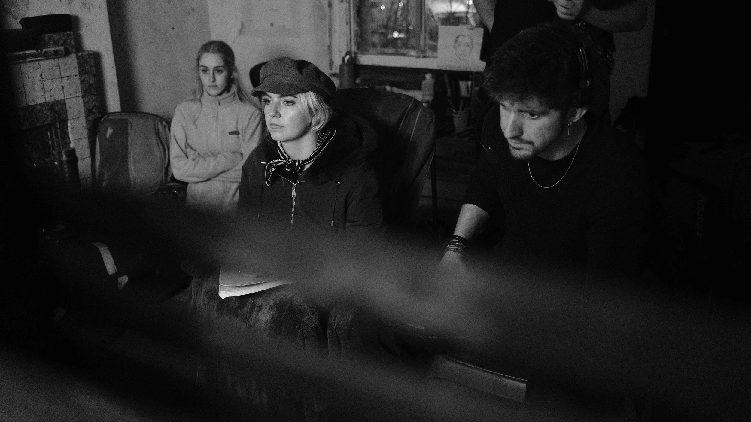 Three people in a dimly lit room; a woman with blonde hair sitting at a desk, a woman with a cap sitting next to her, and a man with headphones on, working on a computer or equipment.