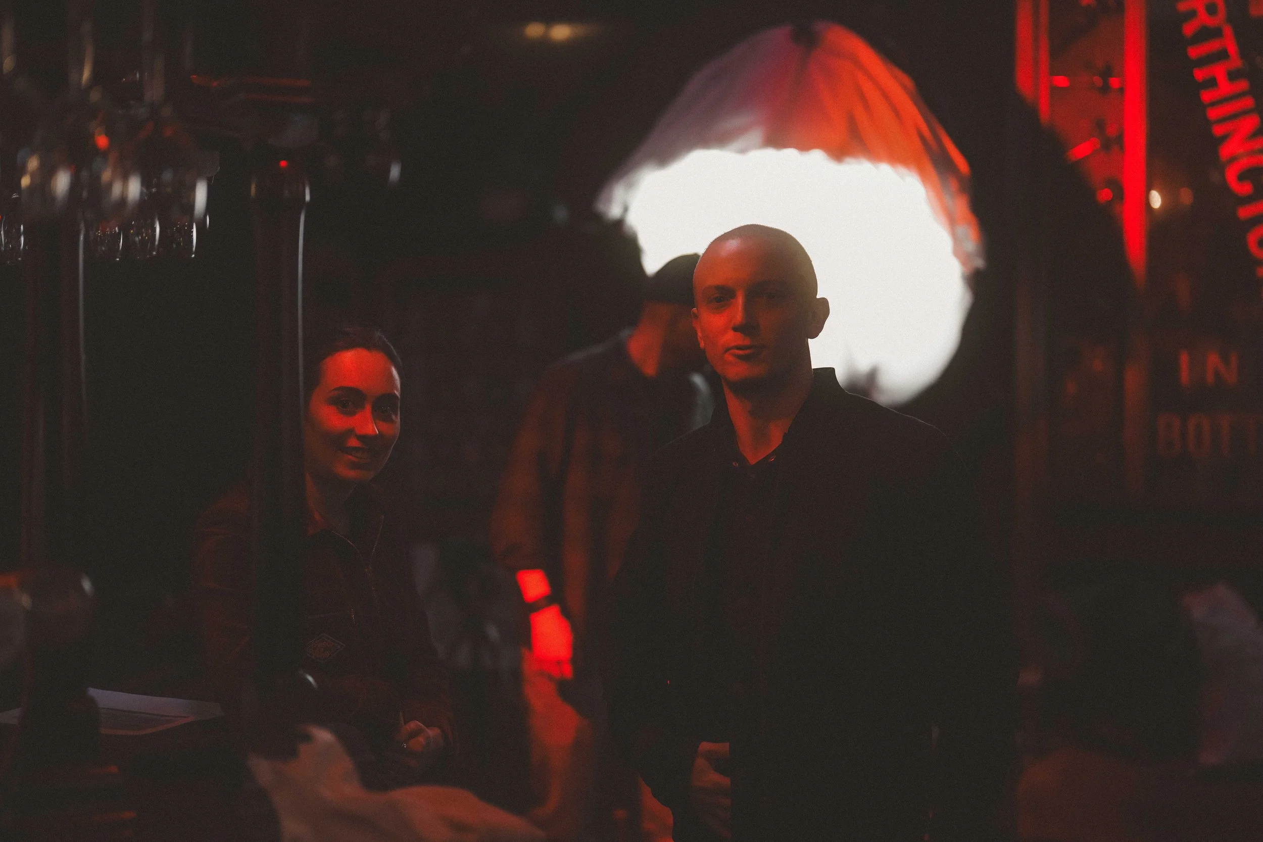 People sitting in a dimly lit indoor space, illuminated by red and orange lighting, with a sign with red letters in the background and a white umbrella overhead.