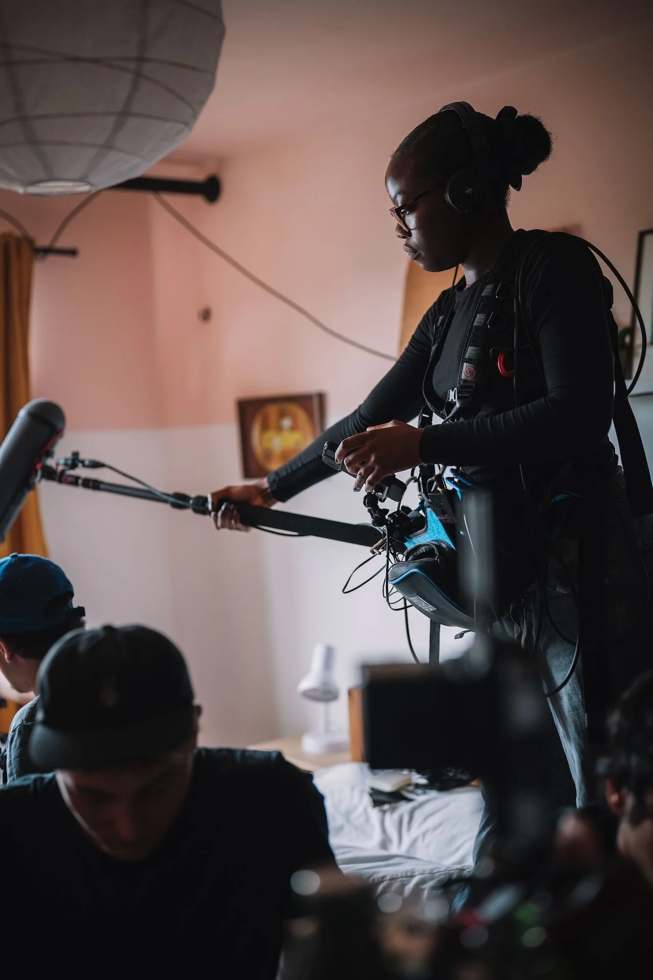 A woman with glasses and a bun hairstyle is wearing headphones and operating a camera mounted on a stabilizer during an indoor event, with several people seated in front of her.