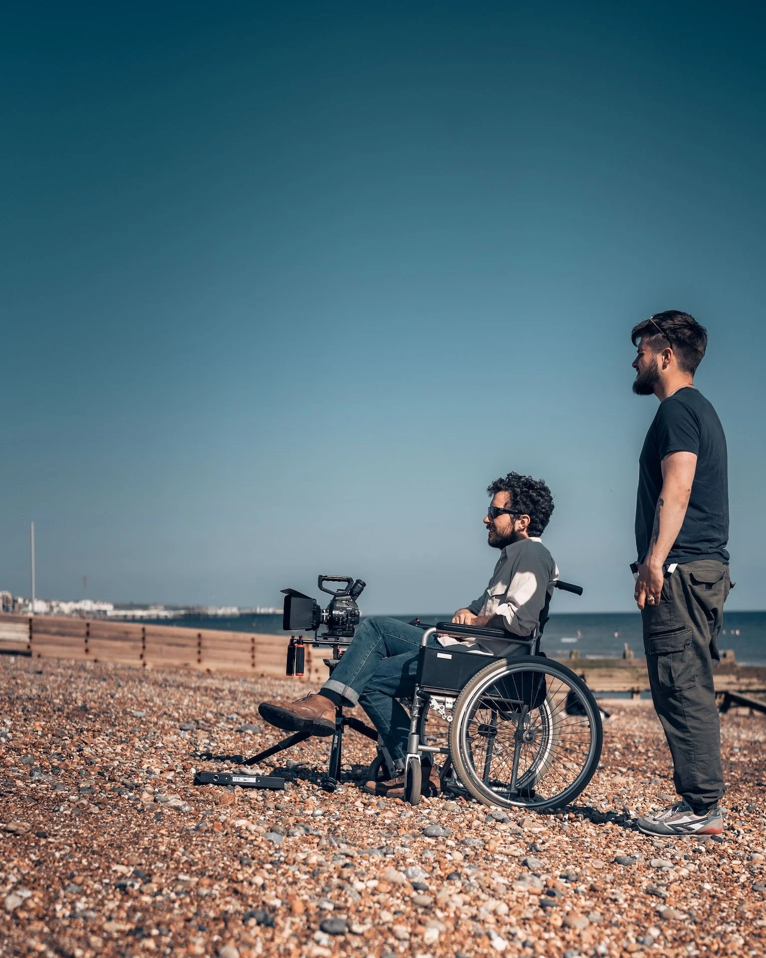 Two men on the beach, one in a wheelchair and the other standing, having a conversation with a calm ocean in the background.