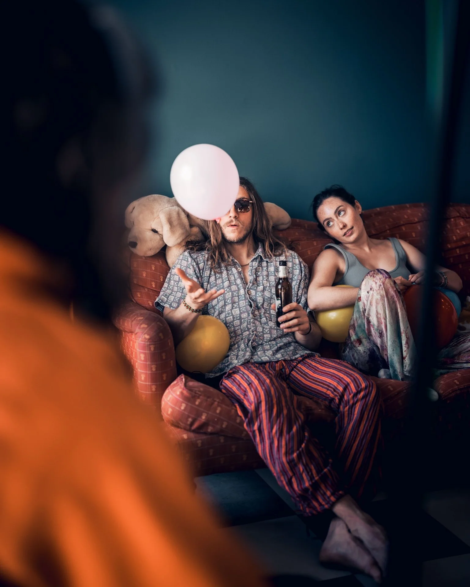 Two people sitting on a red patterned sofa, one holding a bottle, with a teddy bear and balloons around them, in a room with a dark teal wall.