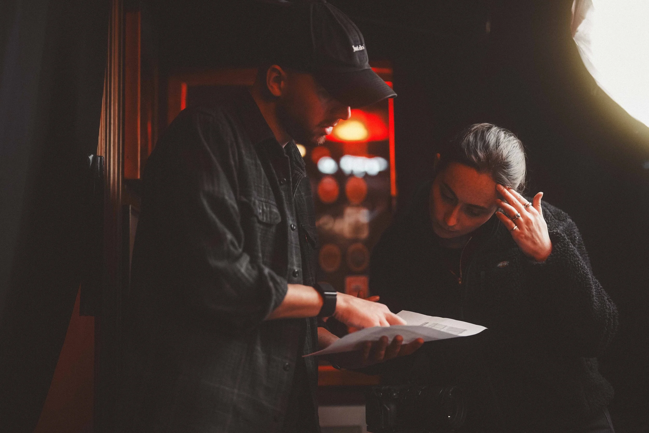 A man and woman playing piano together in a dimly lit indoor setting, with the man wearing a baseball cap and dark jacket, and the woman with her hand on her forehead, appearing to be focused or emotional.