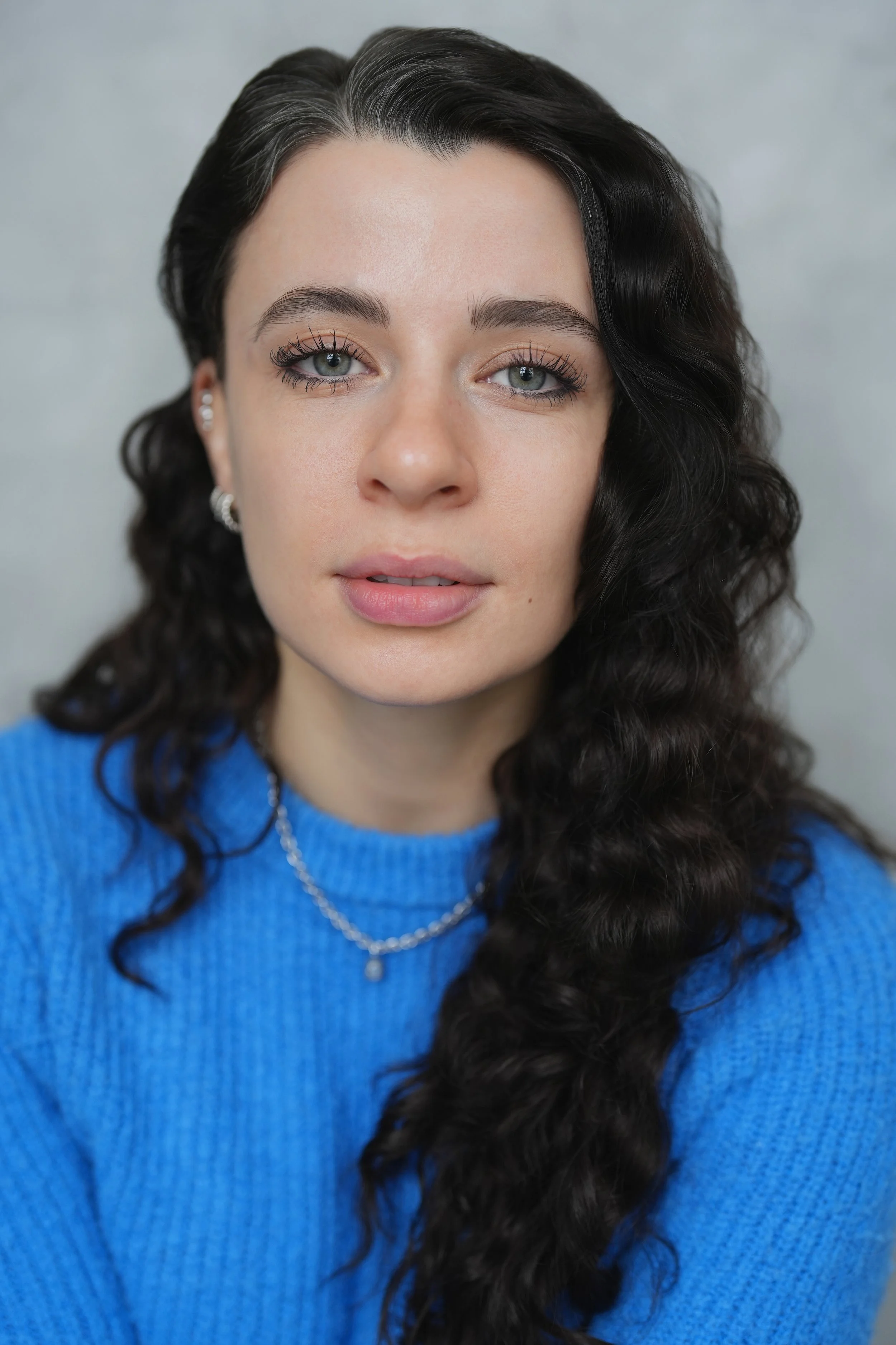 Close-up portrait of a woman with wavy dark hair wearing a blue sweater and jewelry, looking at the camera.
