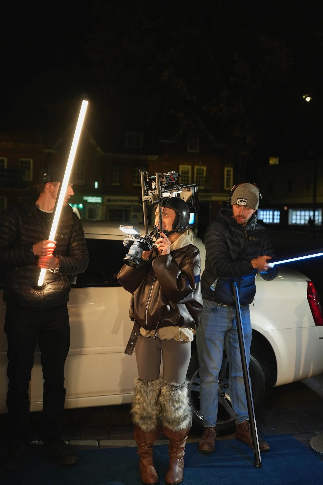 Three crew members working on a film set at night in front of a white vehicle. One person is holding a light stick, another has a camera rig, and the third is reading or inspecting a light panel.
