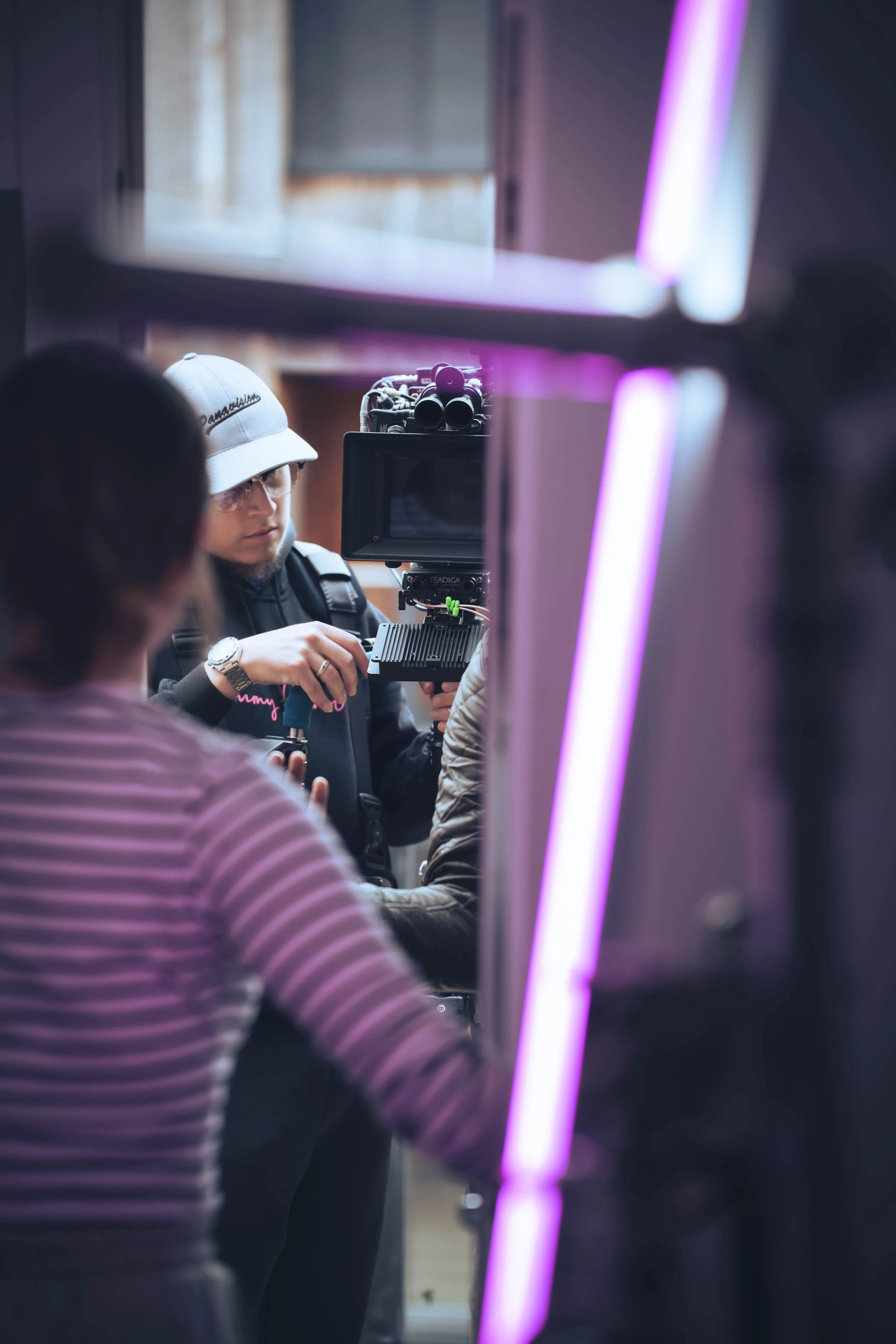 A person wearing glasses, a white cap, and a backpack is operating a camera on a tripod, with a blurred group of people around. There are purple neon lights in the foreground.
