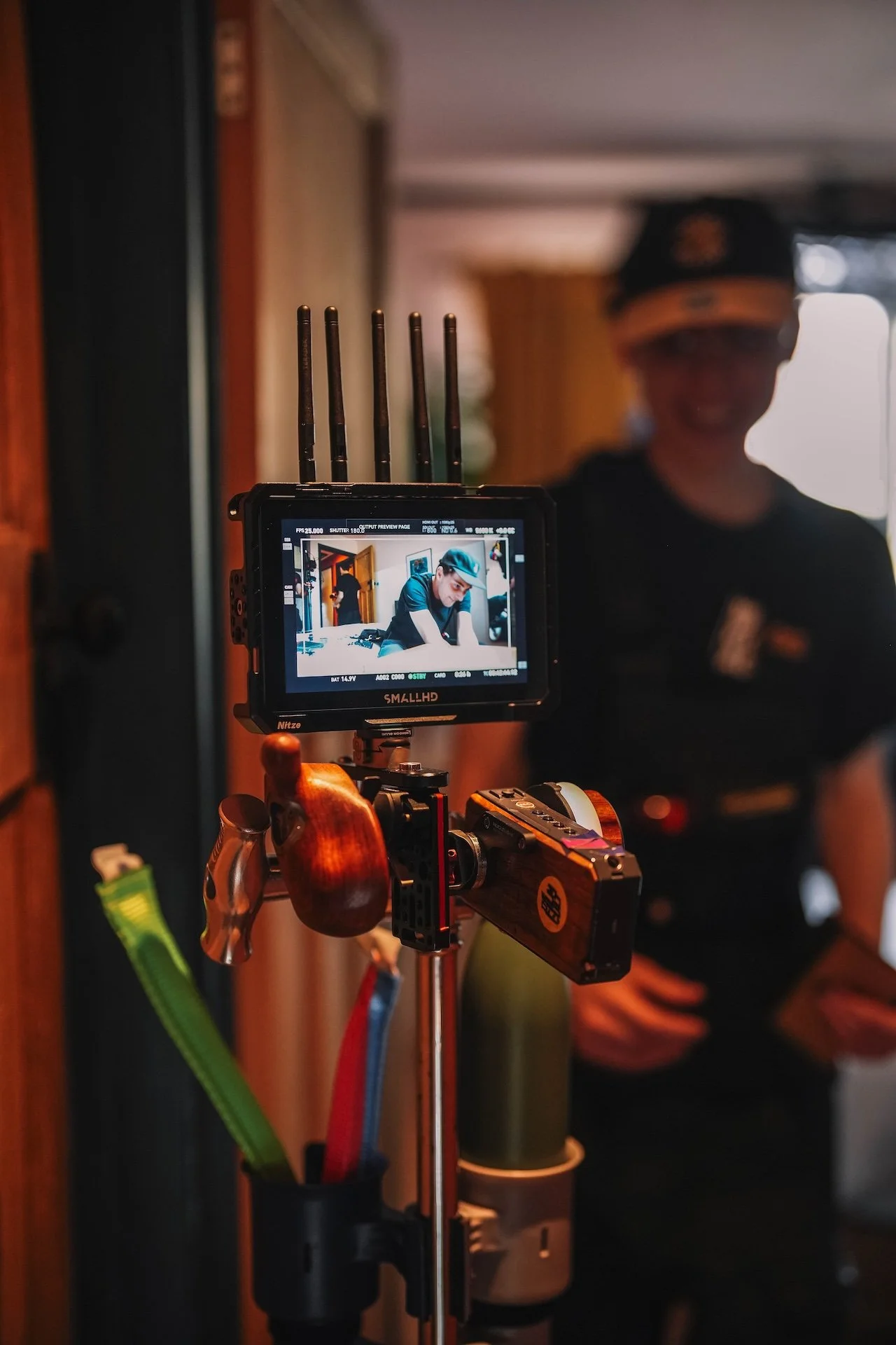 Close-up of a camera monitor on a tripod, capturing a person in a dark shirt and headset, with a person in the background wearing a sporty cap. The setting appears to be indoors.