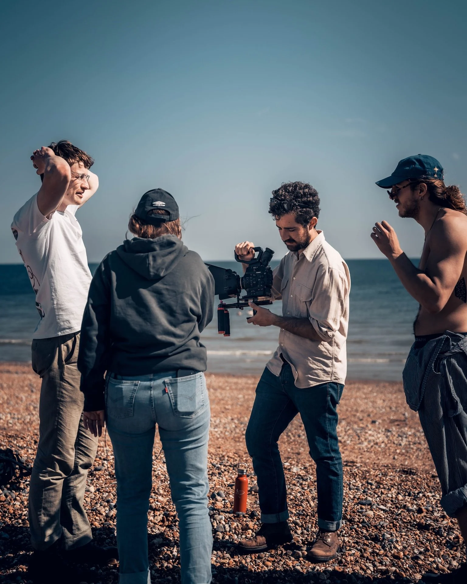 A group of five people standing on a pebble beach, looking at a film camera, with the ocean in the background. They appear to be discussing something, possibly filming or photography, on a clear day.