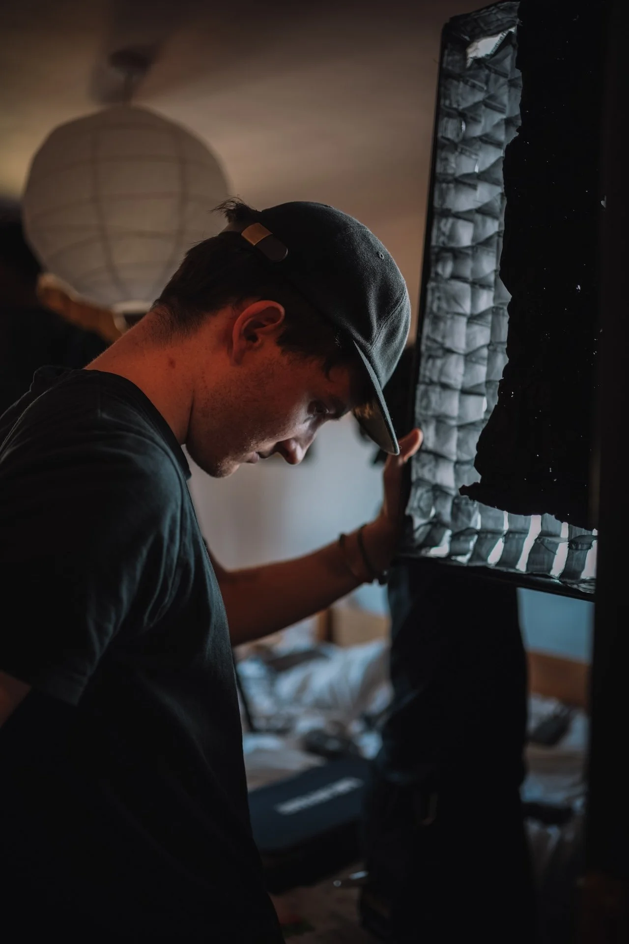 A young man wearing a backwards cap and black T-shirt, looking down, appears to be in a dimly lit room with a loft bed, shelves, and a lamp in the background.