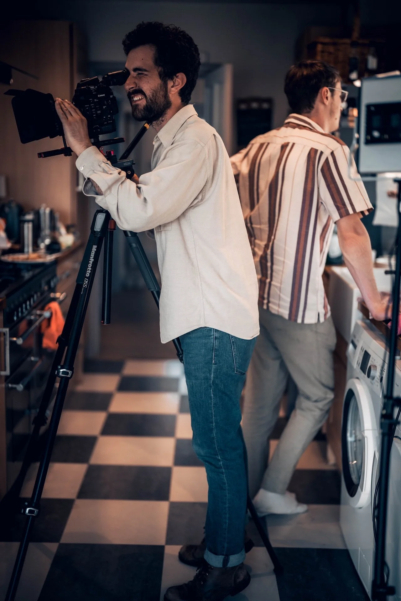 Two men in a kitchen working with electronic and filming equipment. One man looks through a camera mounted on a tripod, while the other is using a laptop near a washing machine.