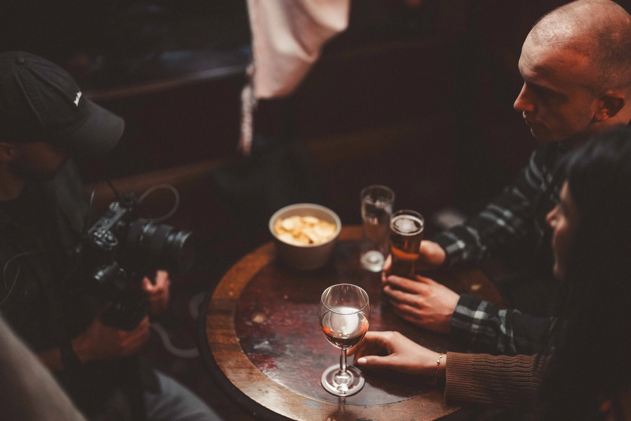 People sitting around a table with drinks and snacks, one person holding a glass of red wine, in a dimly lit setting.