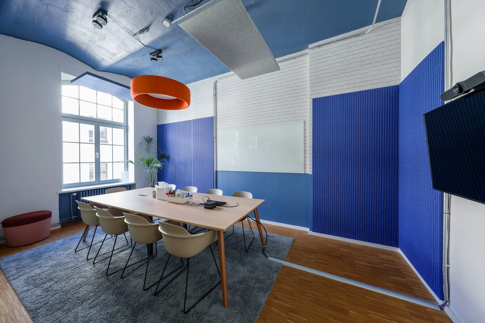 Modern conference room with white walls, blue acoustic wall panels, a large wooden table surrounded by beige chairs, a window with natural light, a whiteboard on the wall, and a flat-screen TV mounted on the wall. There is a large orange ceiling ligh