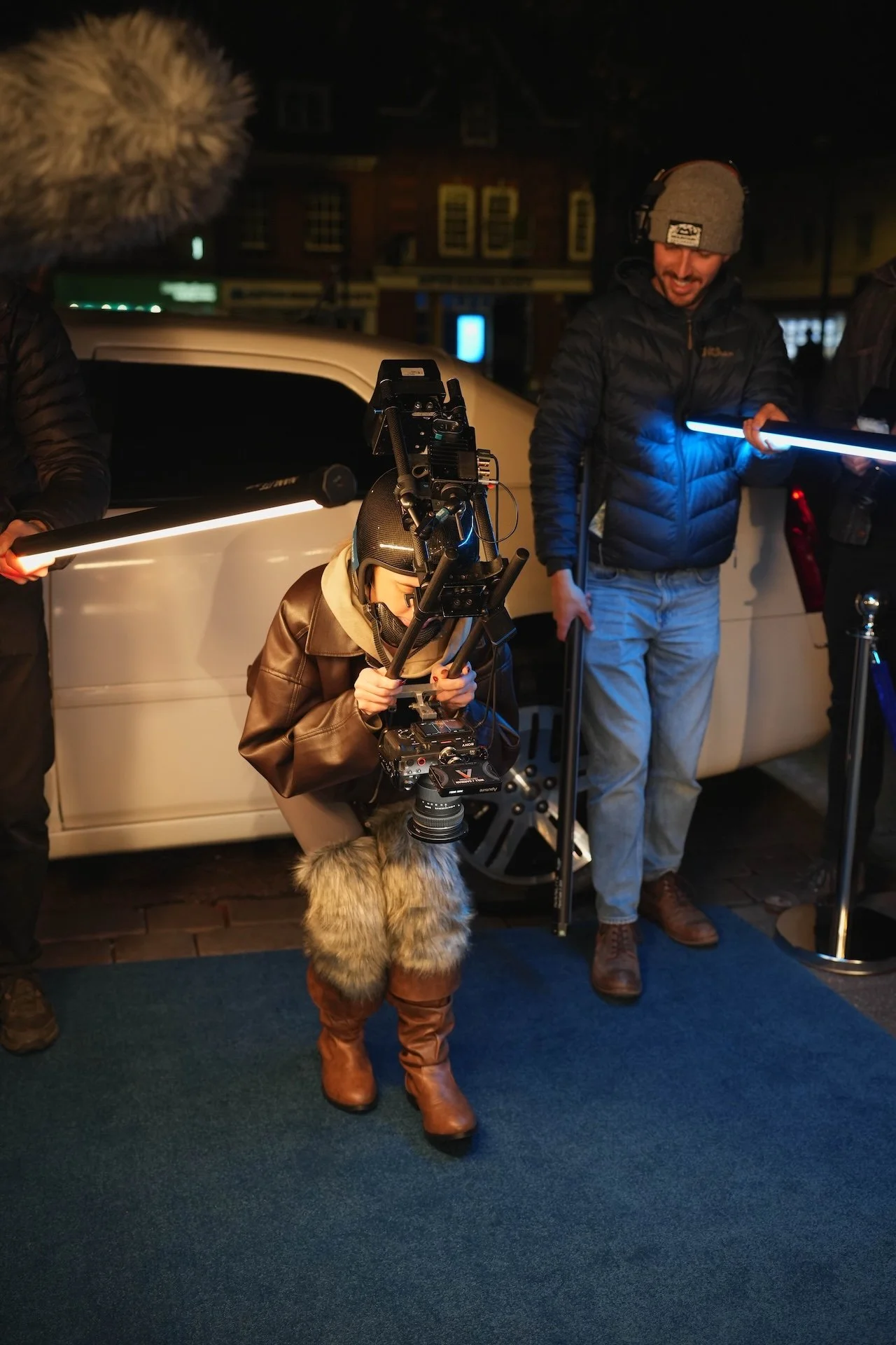 A nighttime scene showing a film crew working outdoors on a street, with a person operating a camera, others holding lighting equipment, and a child in winter clothing standing among them.