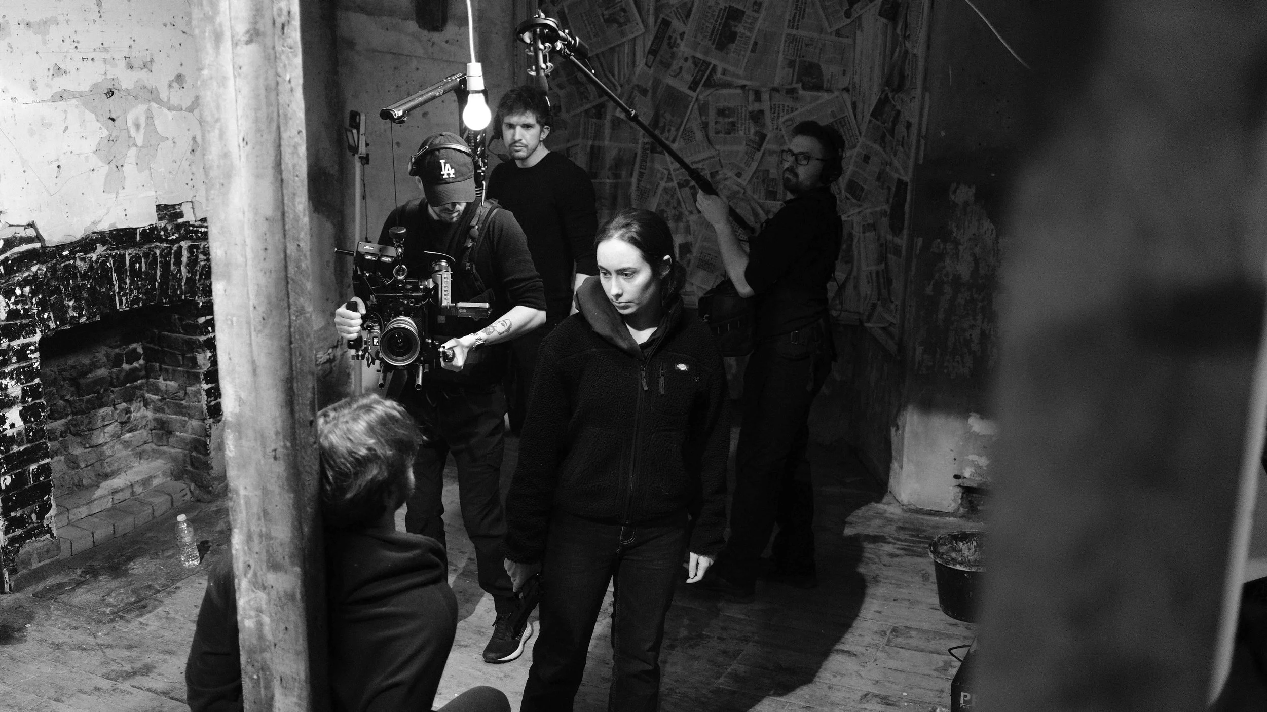 Filmmaking scene with crew members on a set, including a woman in a black jacket holding a gun, and a man operating a camera, with a wall made of newspapers in the background, in black and white.