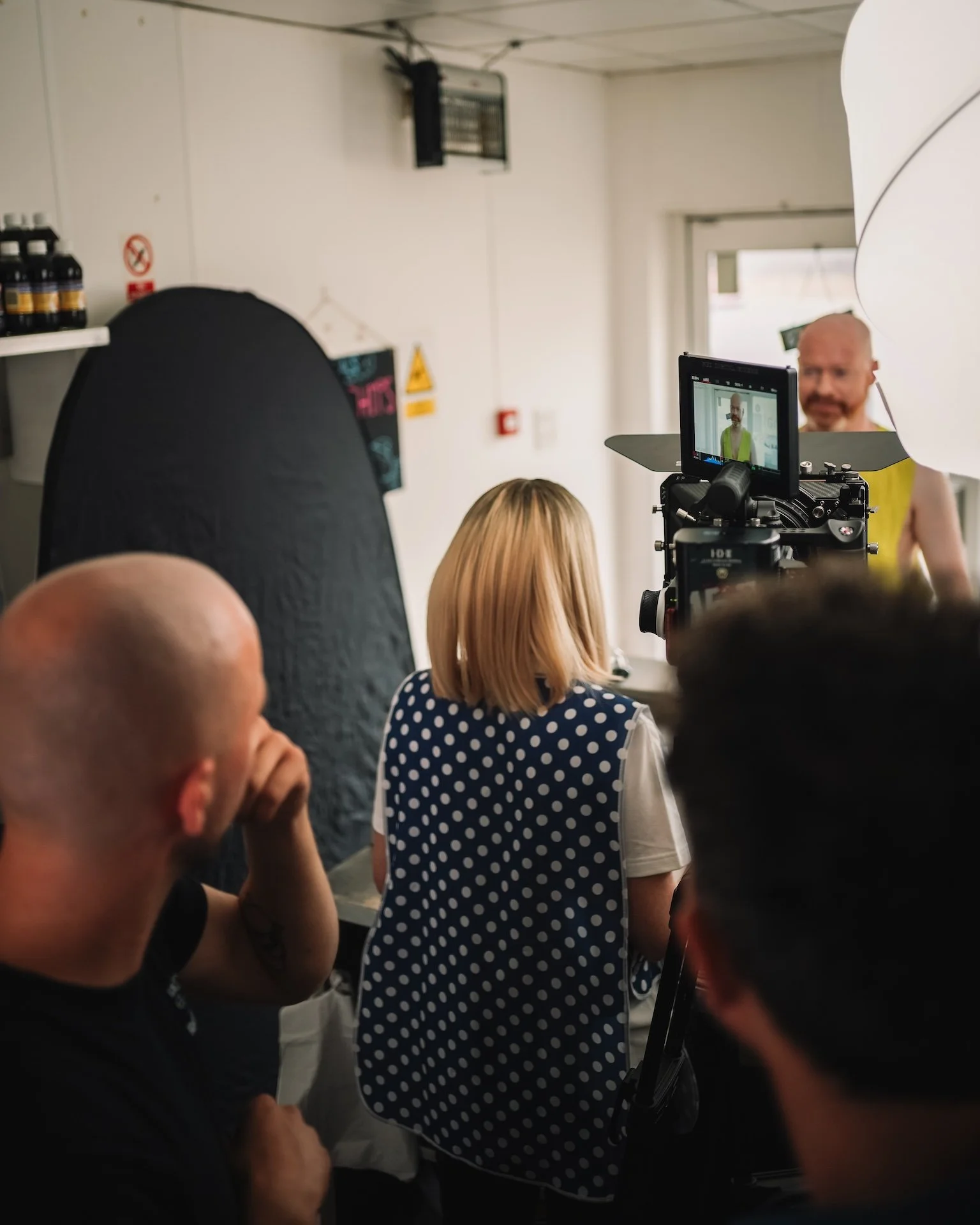 People filming a man with a bald head and beard wearing a yellow shirt during an interview or recording in an indoor setting.