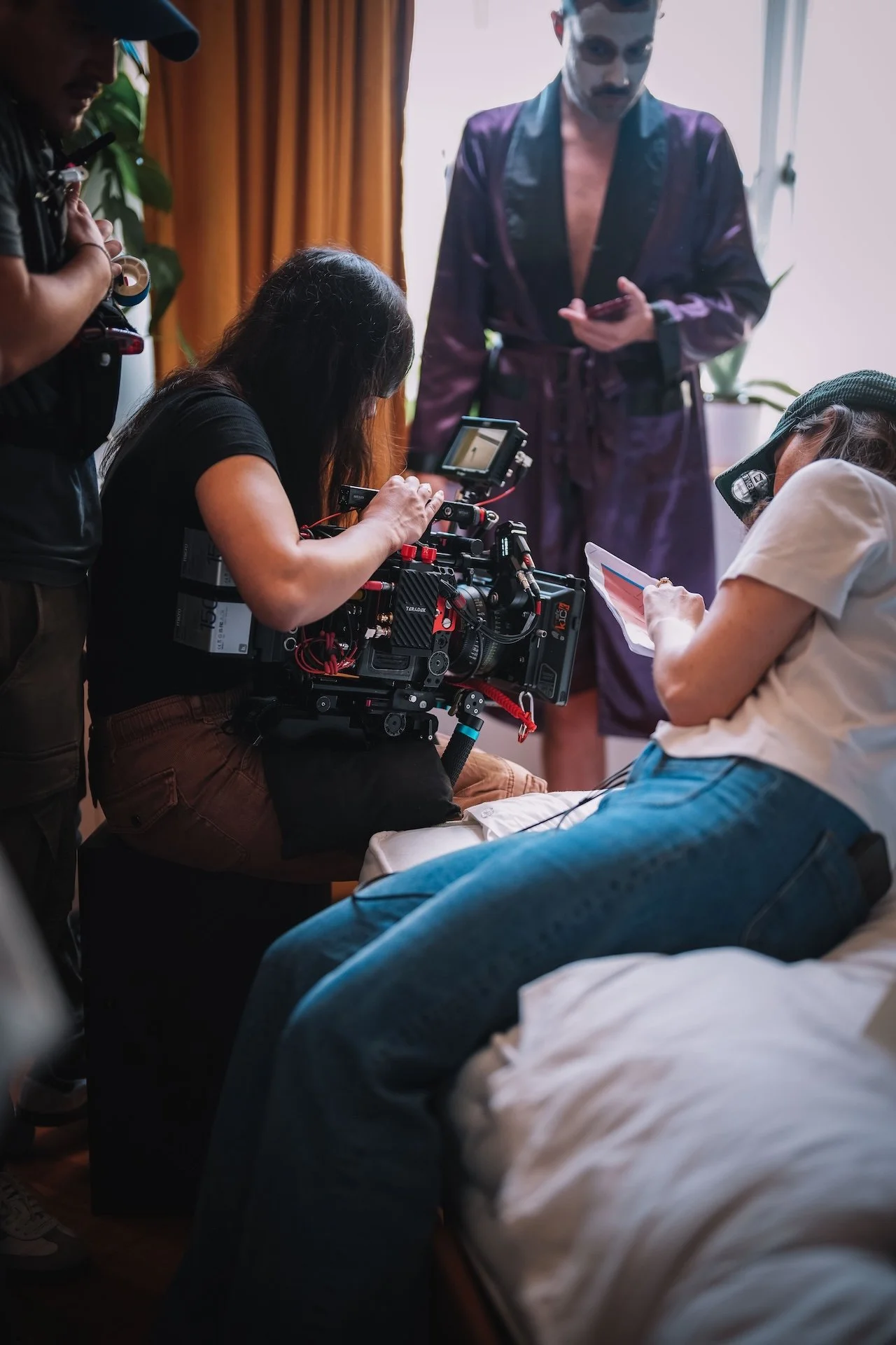 Behind the scenes of a film shoot showing a film crew filming a scene with a person lying in bed, a woman holding a script, and a man in a robe standing nearby.