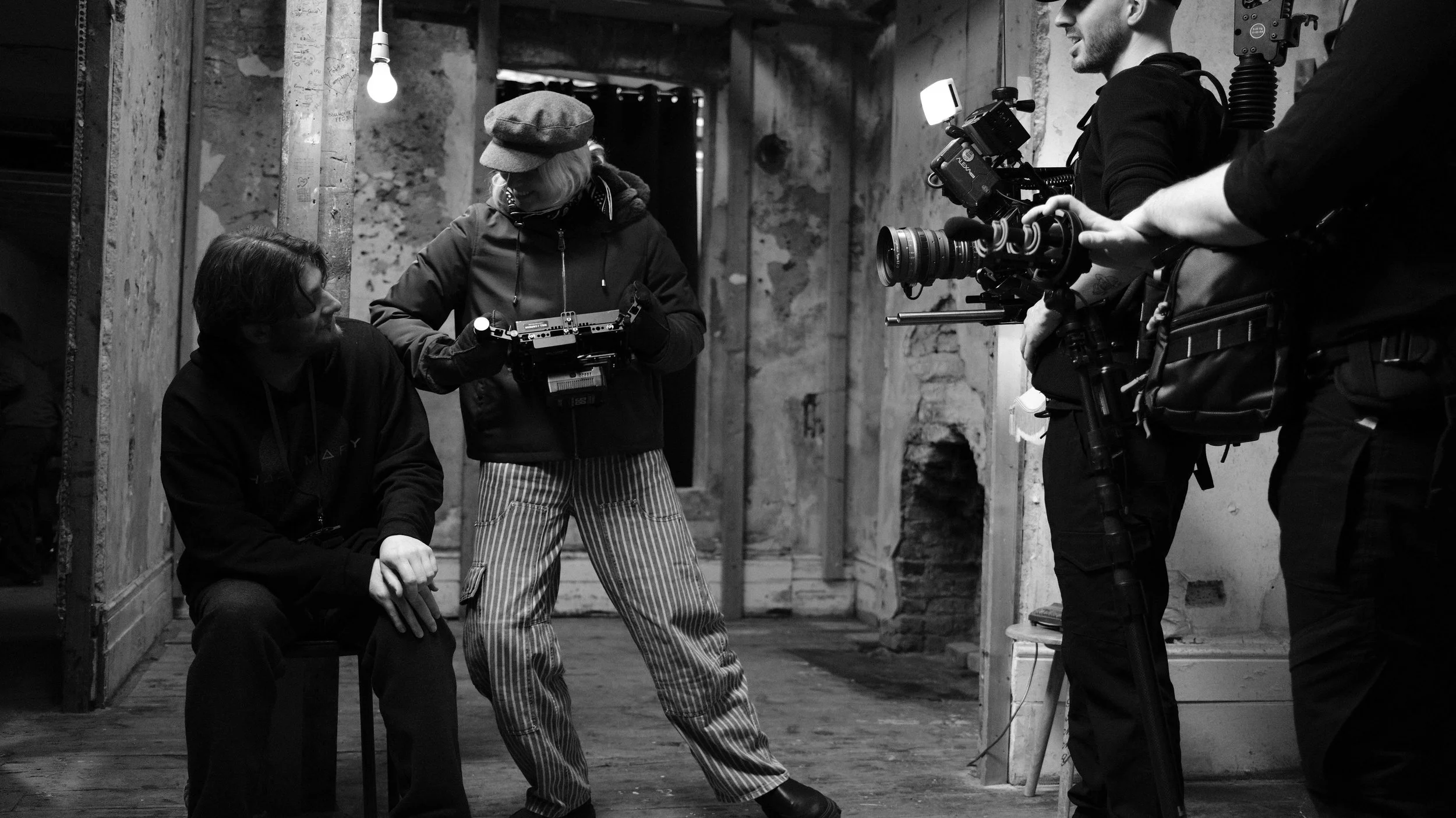 Black and white photo of a film crew on set. A person is sitting on a bench, and a woman is standing and holding a camera, possibly directing. Two crew members are operating a large professional camera, filming the scene.