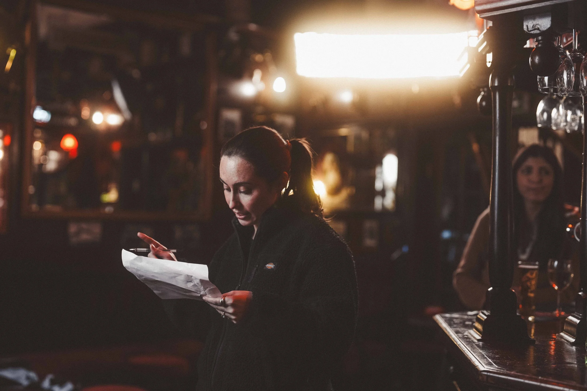 A woman with dark hair tied in a ponytail, wearing a dark jacket, is looking at and possibly using her phone while holding a paper. She is in a dimly lit indoor setting, possibly a bar or pub, with blurred background lights and glasses hanging upside