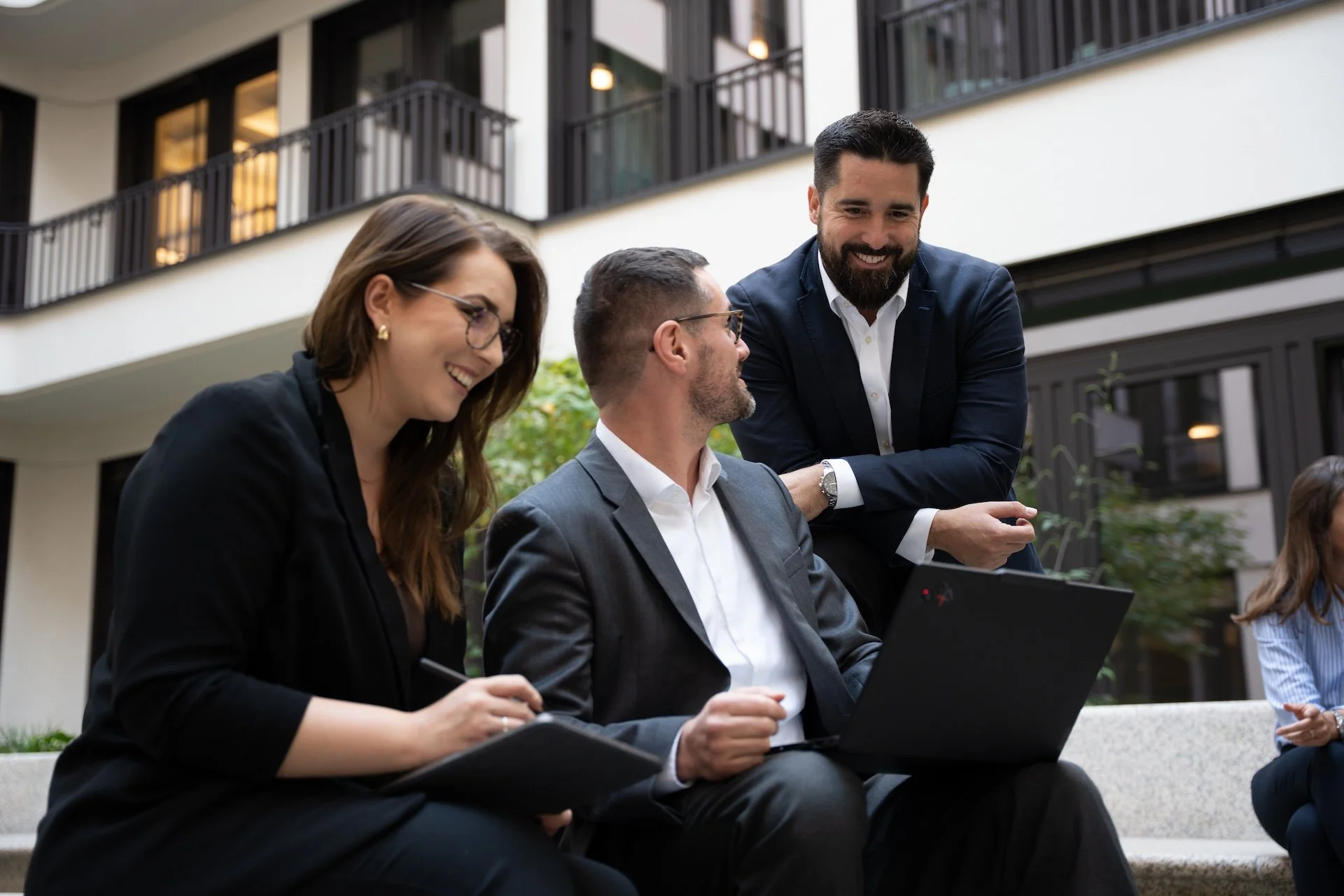 Three business professionals, two men and one woman, engaged in a discussion outdoors near a modern office building. The woman is writing on a notepad, and the men are listening and smiling. One man has a laptop open on his lap.