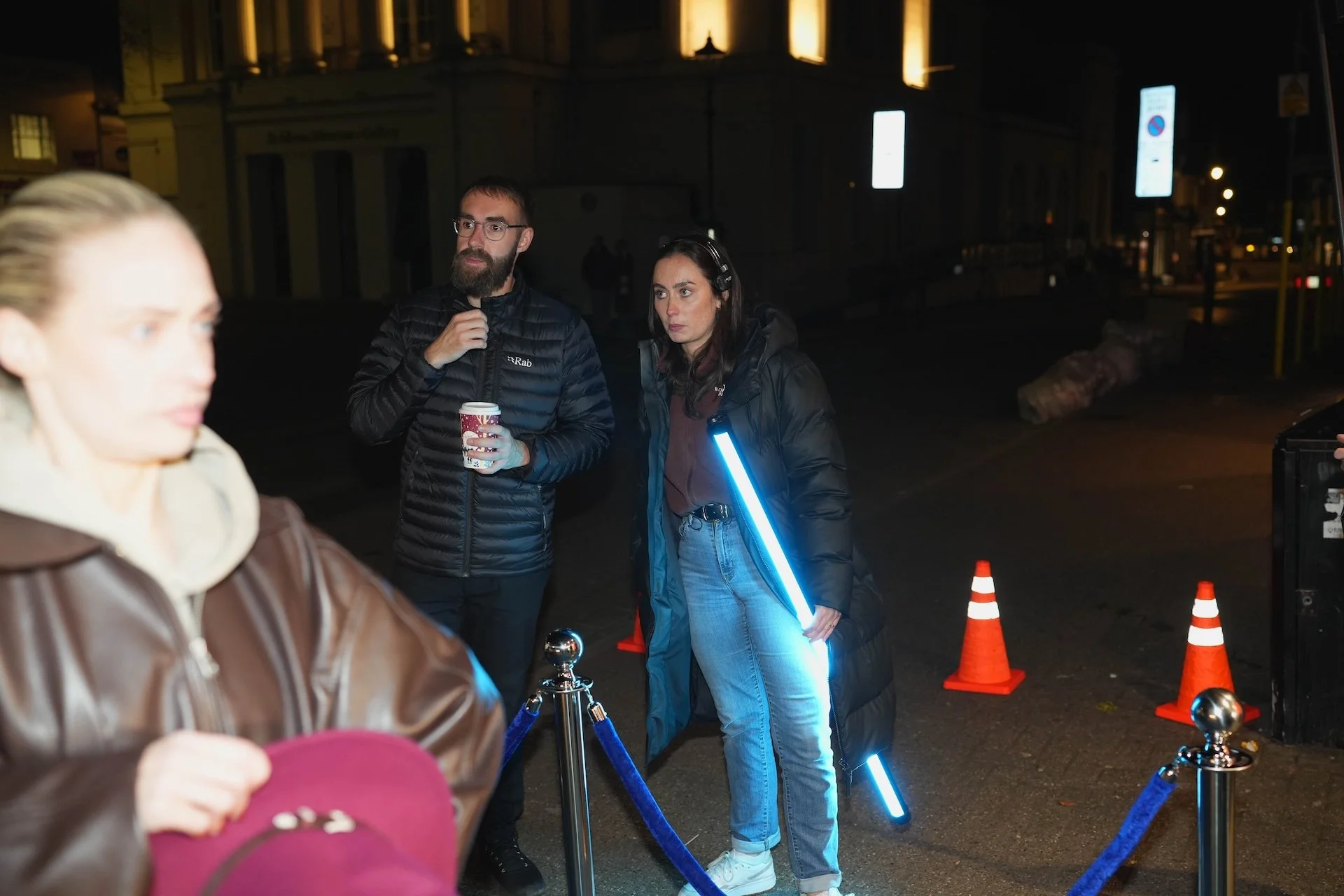Three people standing outdoors at night behind a blue rope barrier, illuminated by a glowing blue stick, near orange traffic cones. One woman in the foreground is partially visible, wearing a brown leather jacket and holding a pink hat. In the backgr