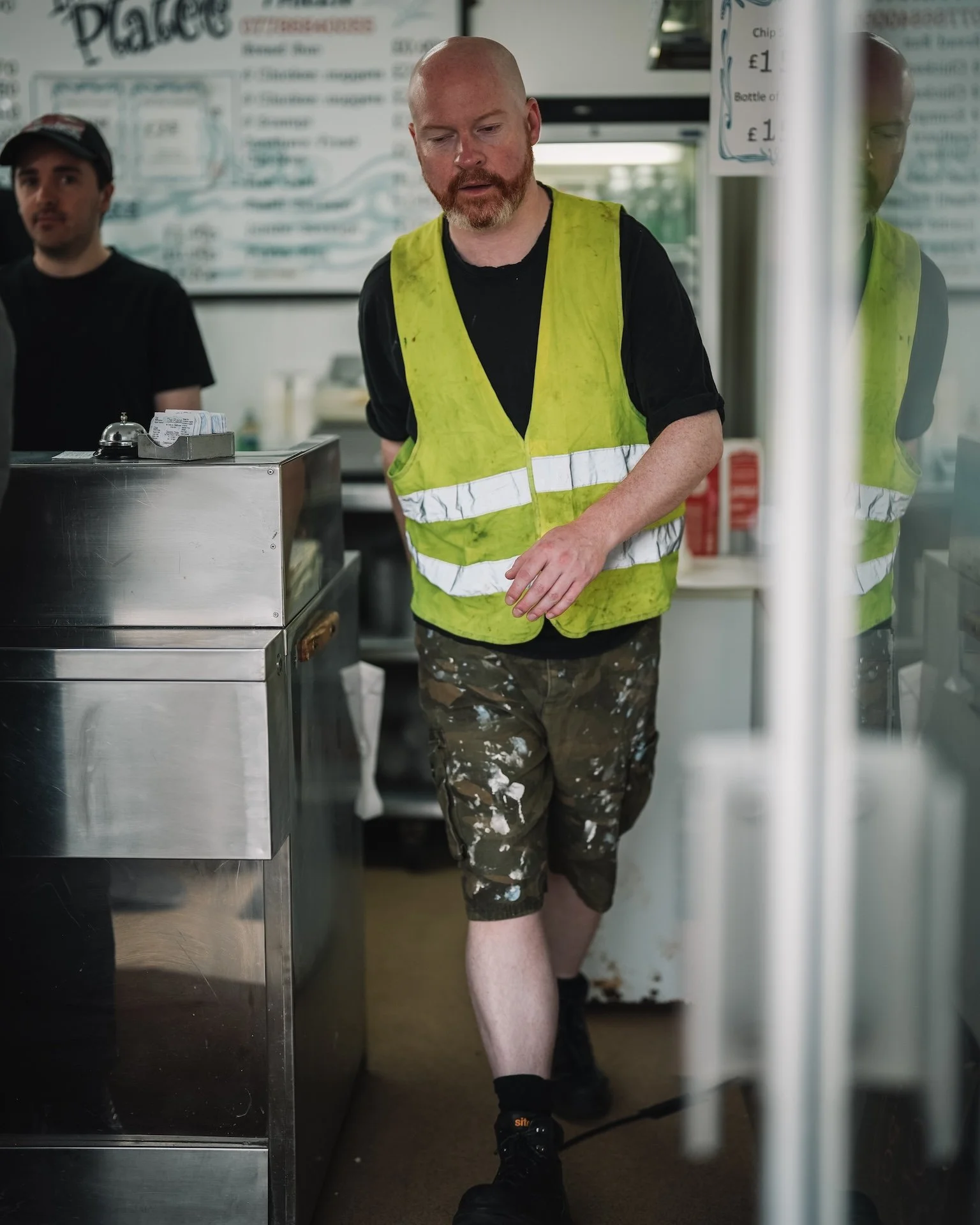 A man with a shaved head, beard, and light skin wearing a yellow safety vest, black shirt, and camouflage shorts, walking through a food establishment door.