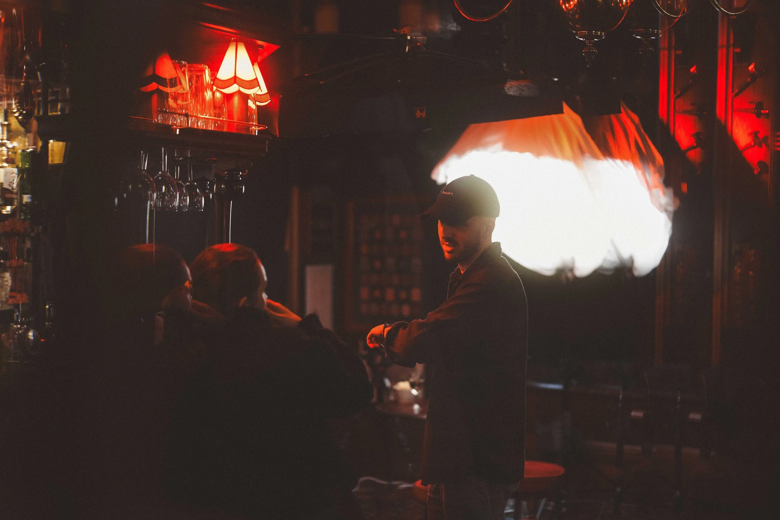 A bartender or server wearing a cap behind a bar illuminated by red and warm lighting, with shelves of liquor bottles and hanging glasses in the background.