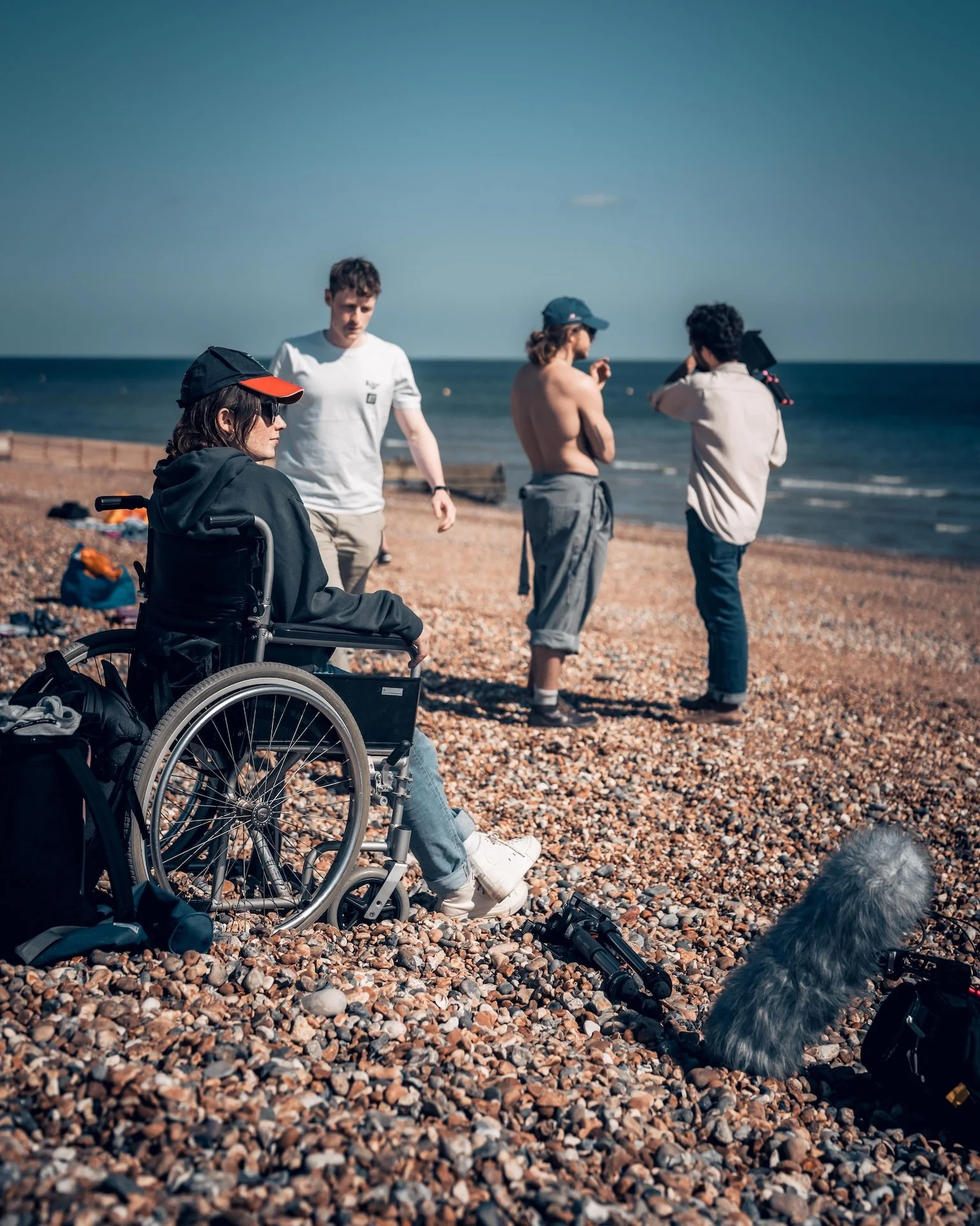 Group of four people on a pebble beach, with one in a wheelchair, and a dog, overlooking the ocean, some people are talking and one is looking through binoculars.