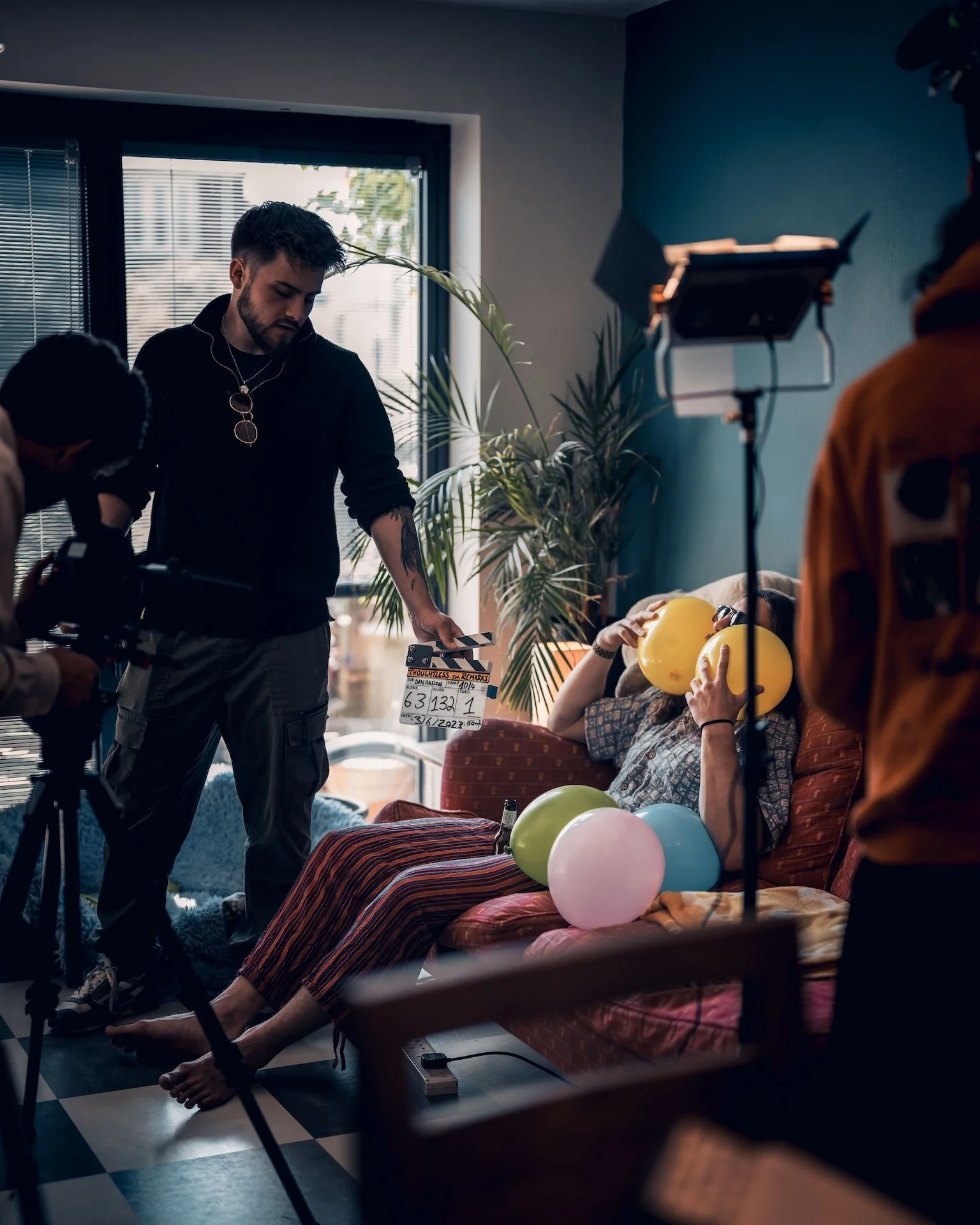 Filming a scene with a woman sitting on a sofa, holding yellow balloons to her face, surrounded by colorful balloons, while crew members film her on a set with natural light coming through a window and film equipment visible.