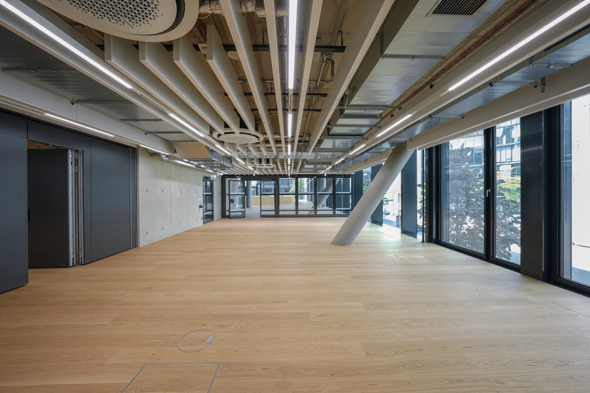 Empty modern interior space with wooden flooring, large windows, and exposed ceiling with white structural beams and linear lighting.