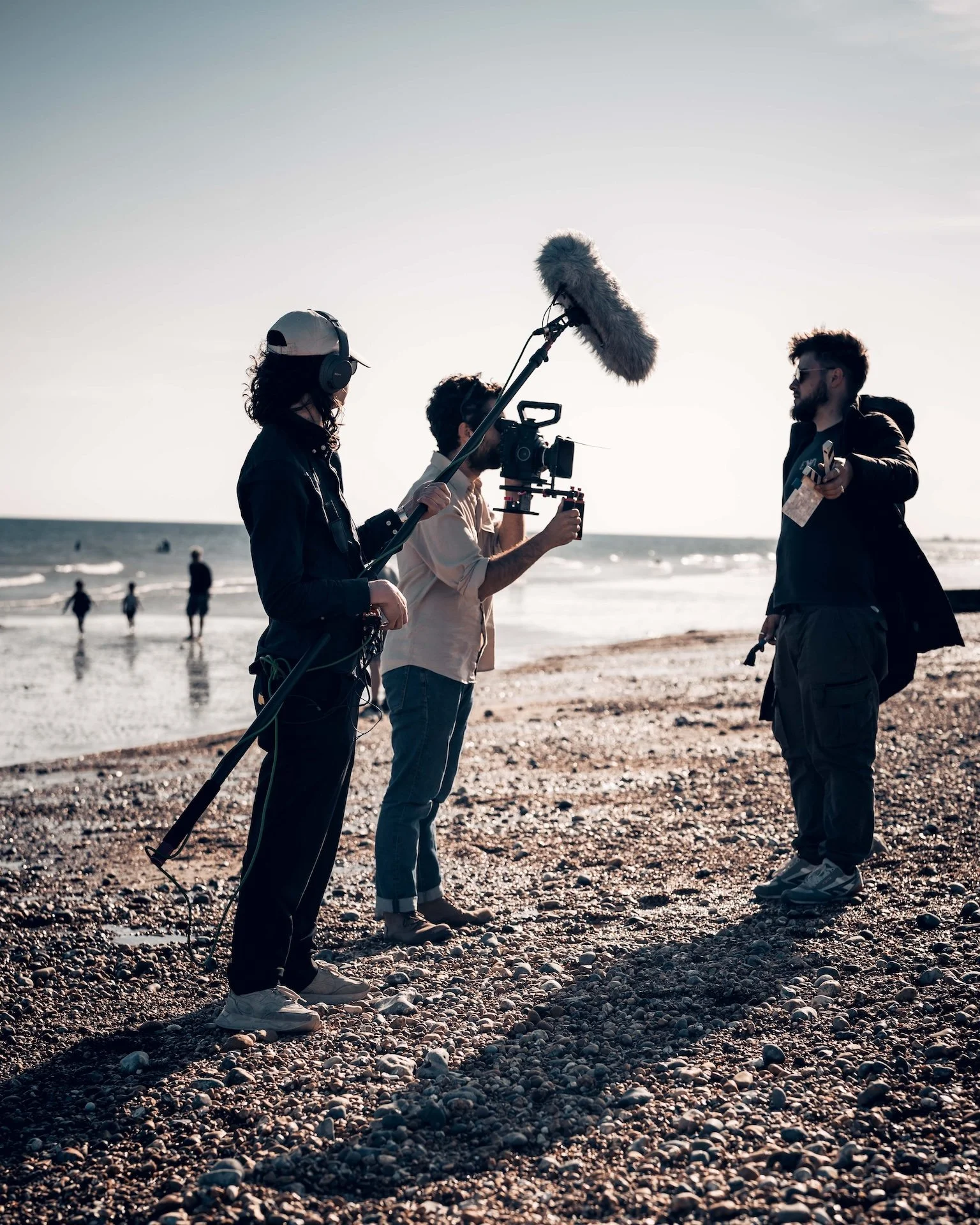 Filmmakers filming on a pebbled beach at sunset with cameras and sound equipment, a man holding a clapperboard, and the ocean in the background.