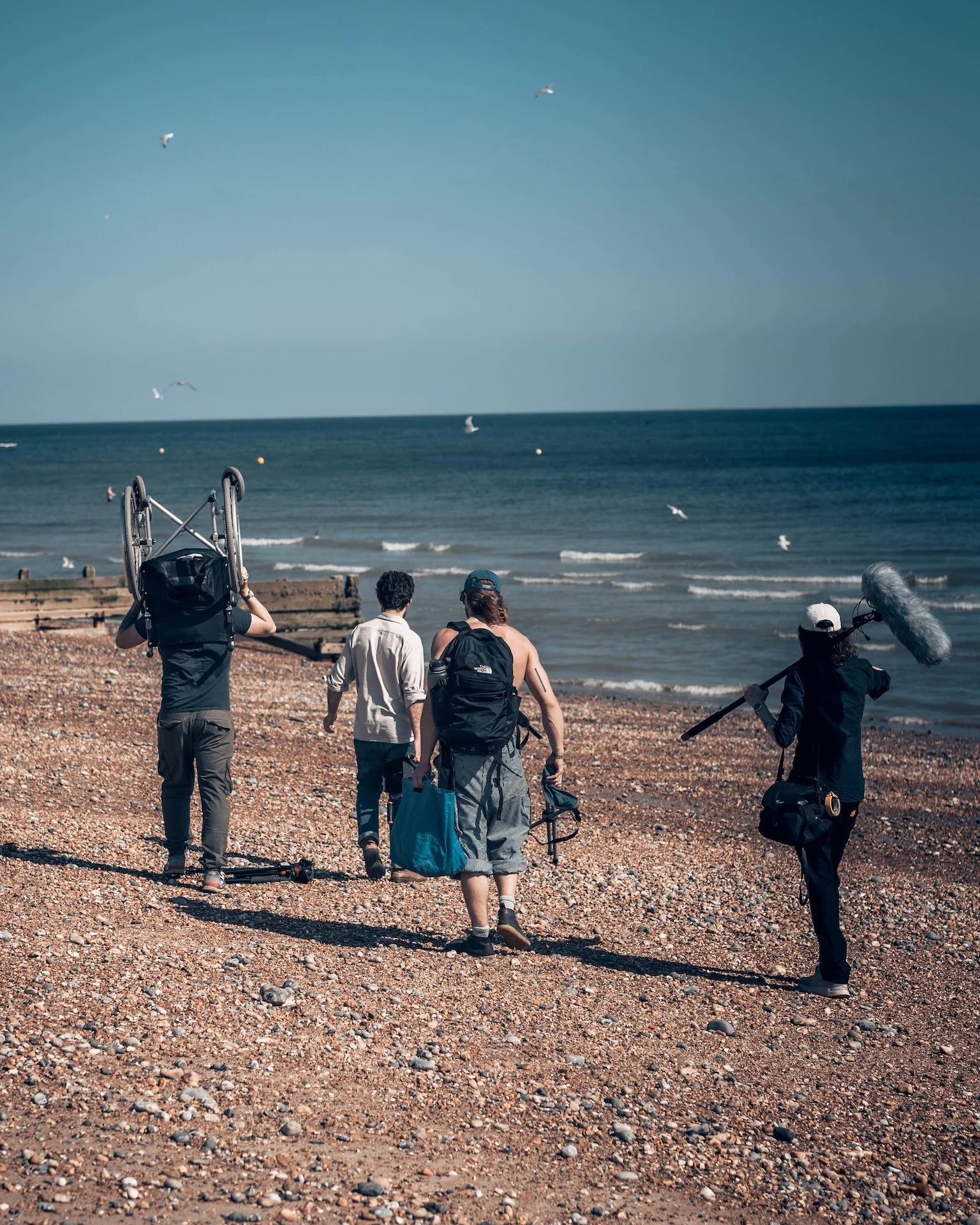 Group of five people walking on a pebble beach towards the ocean, with seagulls flying overhead. One person is carrying a folded wheelchair, and another is holding a boom microphone.