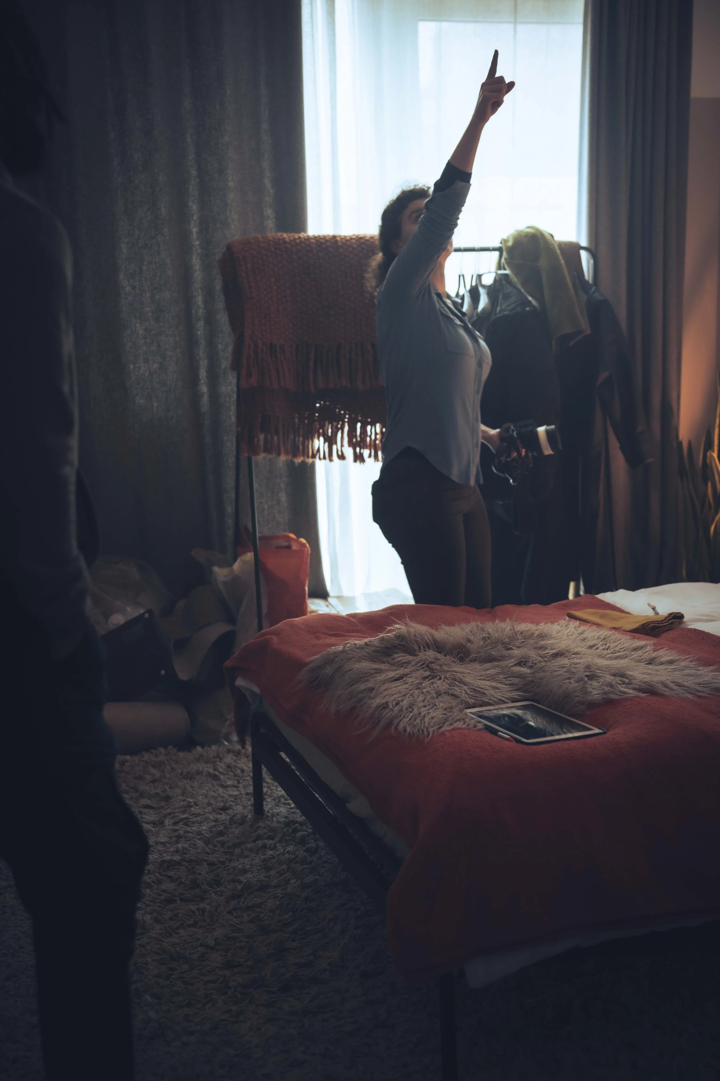 A woman stands in a bedroom with her arm raised pointing upward. She is near a clothing rack with jackets and a blanket draped over it. The room has a window with sheer curtains, and a bed with a furry blanket and an electronic device on it. The ligh