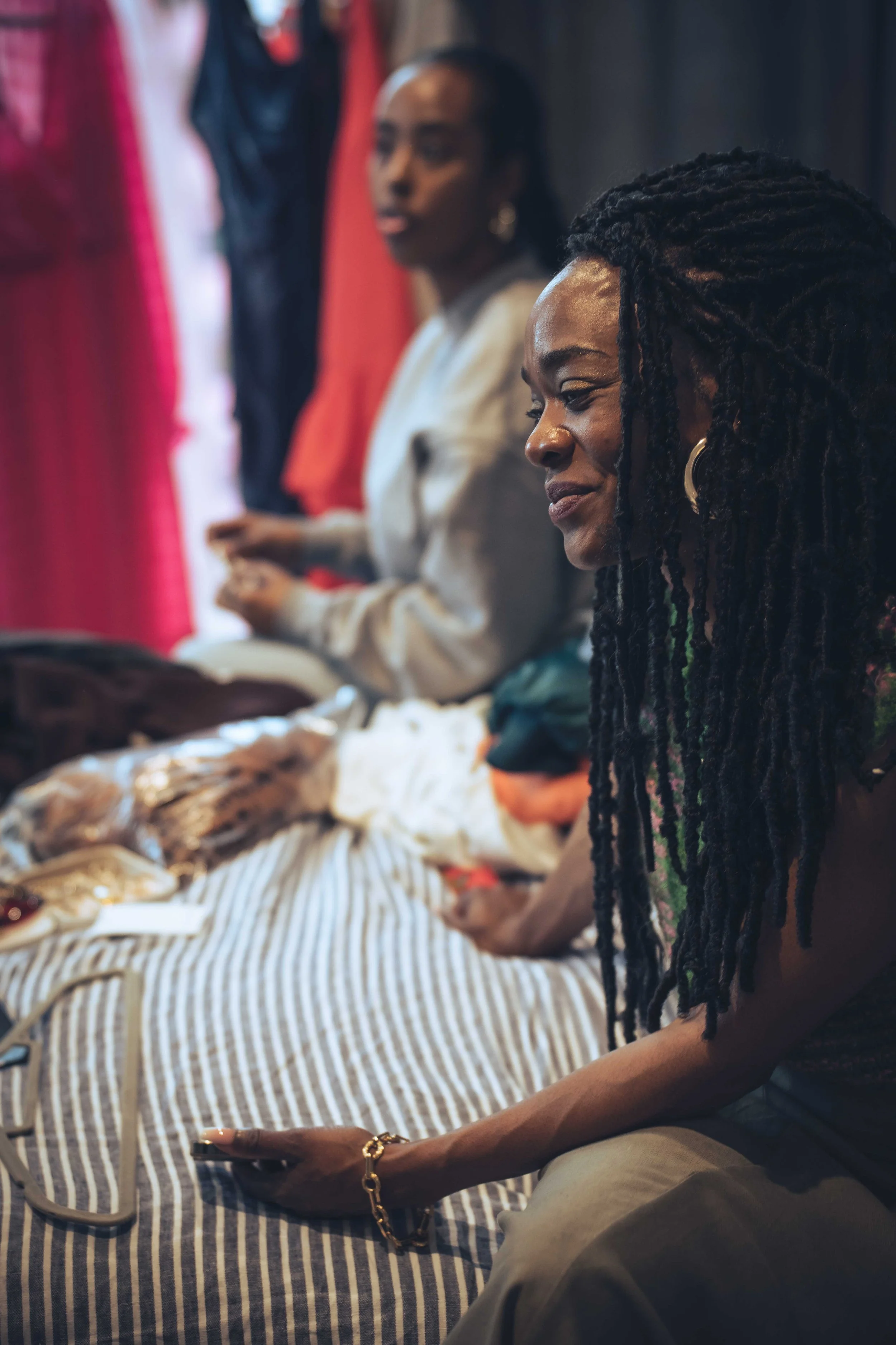 Two women seated at a table with fabric and jewelry, one using a phone and the other looking slightly to the side, with colorful clothing hanging in the background.