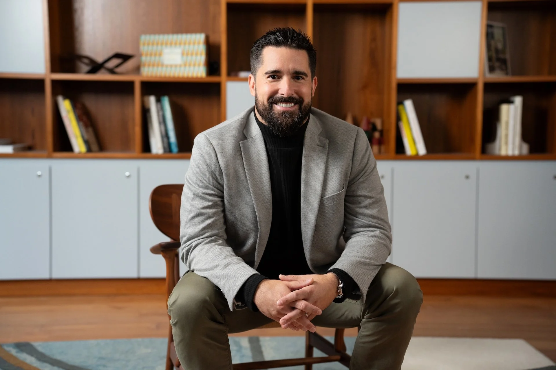A man sitting on a wooden chair, smiling, with a bookcase behind him filled with books and decorative items in a modern office setting.