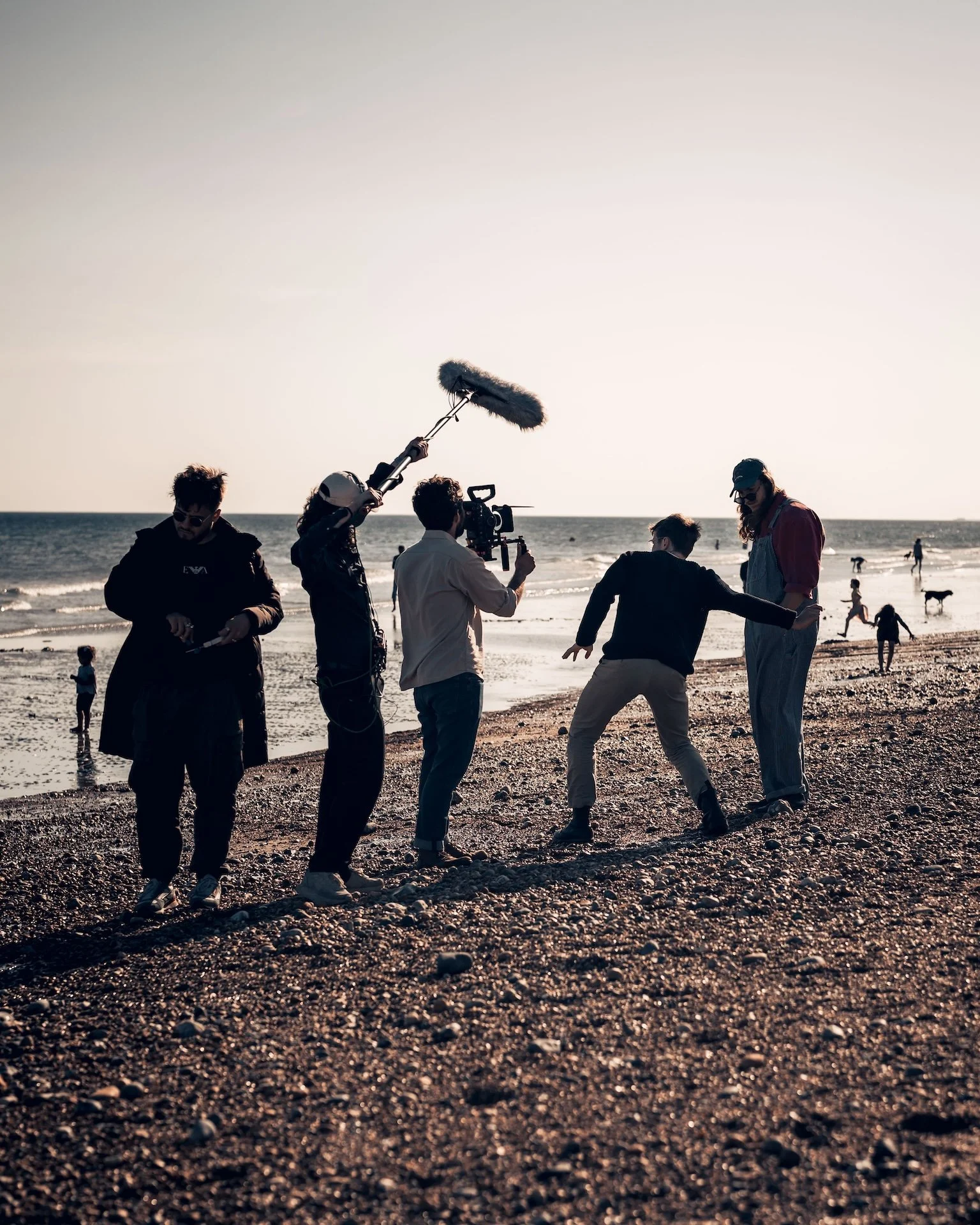 Filmmakers filming a scene on a beach at sunset, with people and dogs in the background.