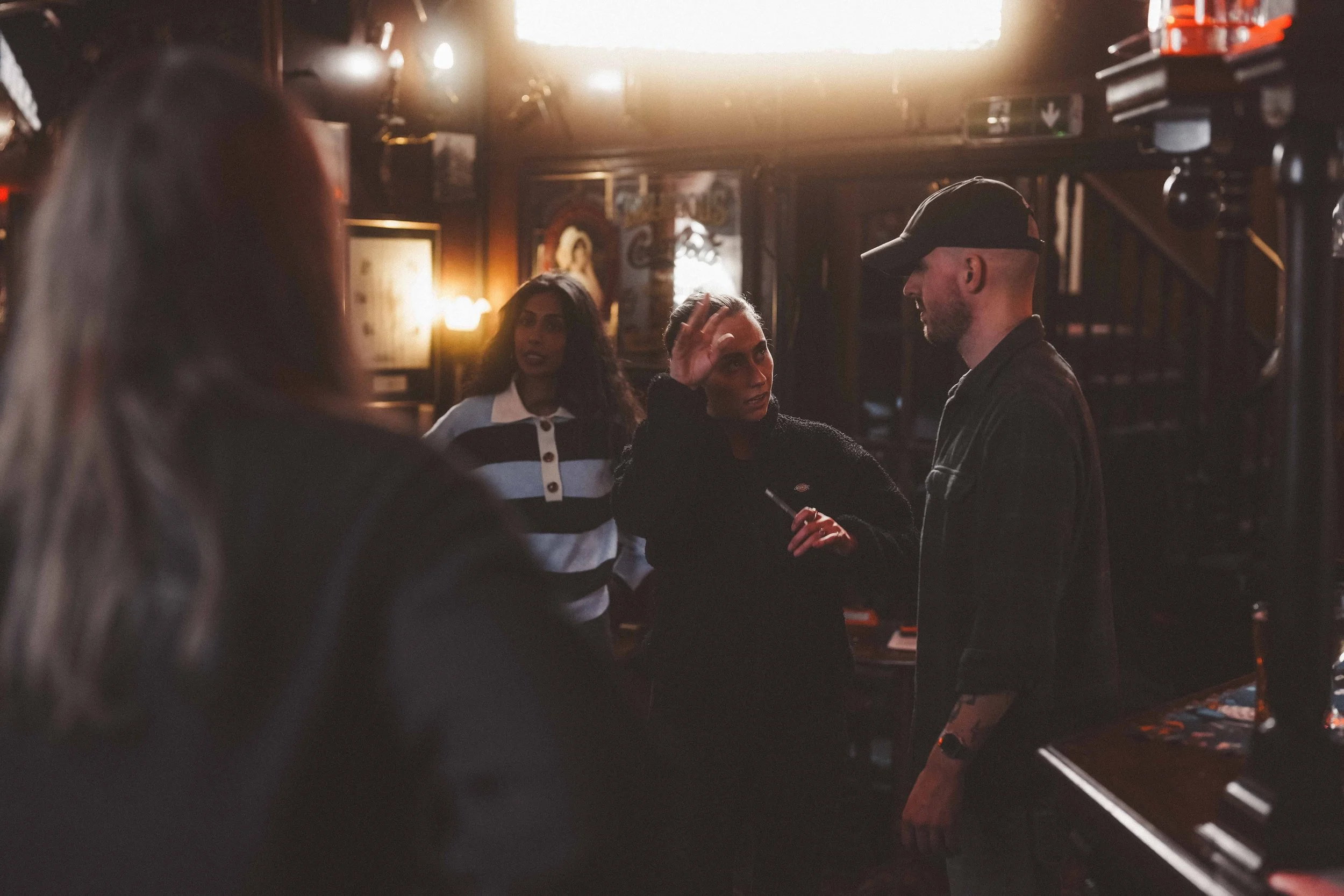 Three people are having a conversation in a dimly lit bar. One woman is talking and gesturing with her hand, while another woman and a man are listening.