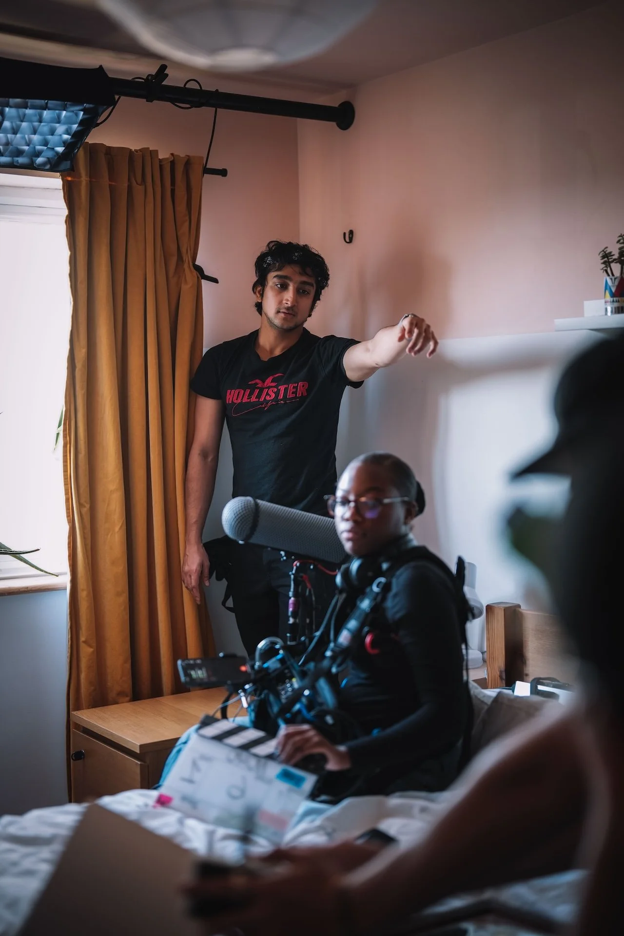A young man in a black T-shirt with red writing stands in a room, reaching out with his right arm. In the foreground, a woman with glasses and dark clothing is sitting in bed with medical equipment and papers, while a person is partially visible and 