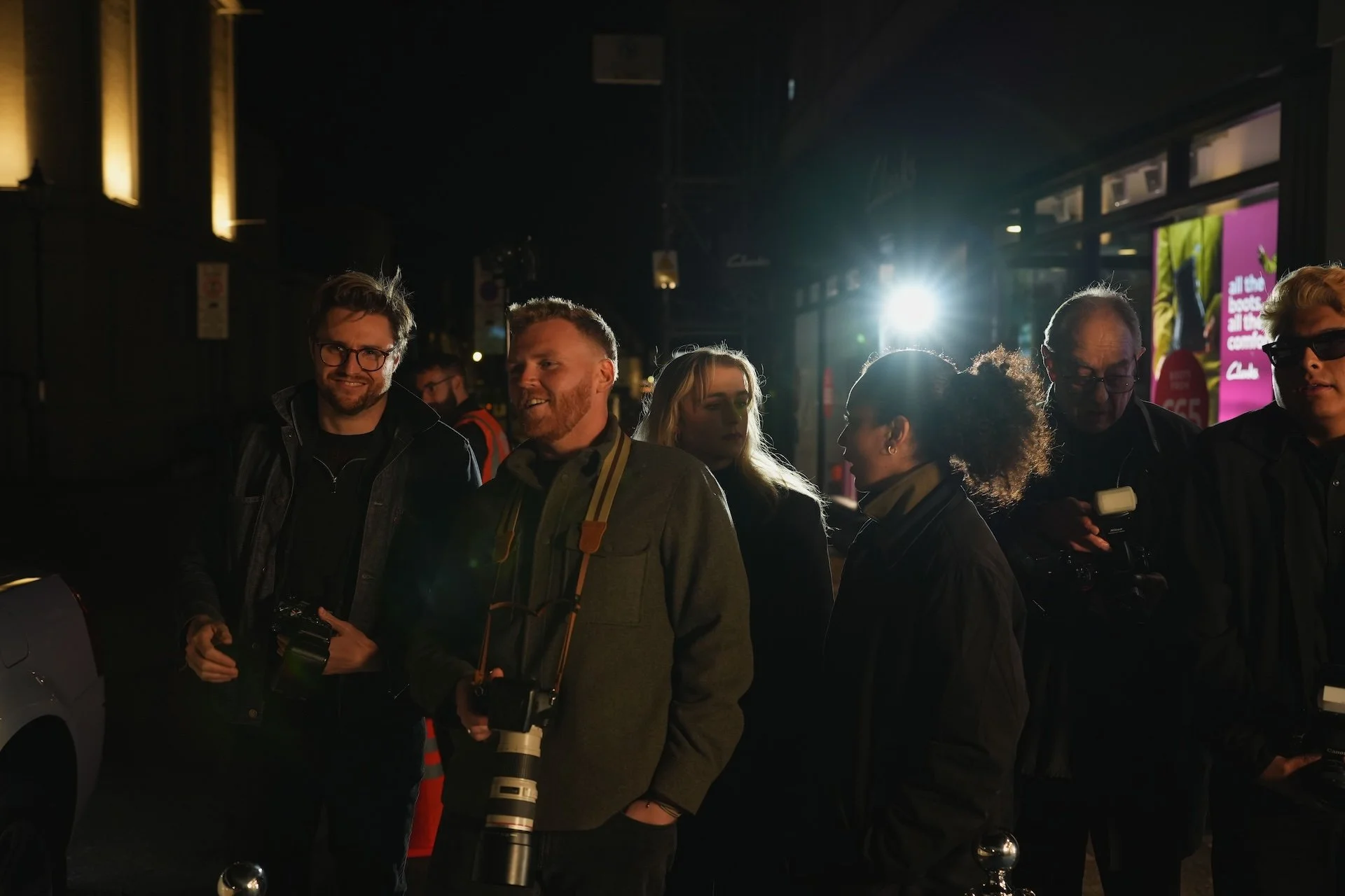 Group of people standing outdoors at night, some holding cameras, illuminated by bright light behind them.