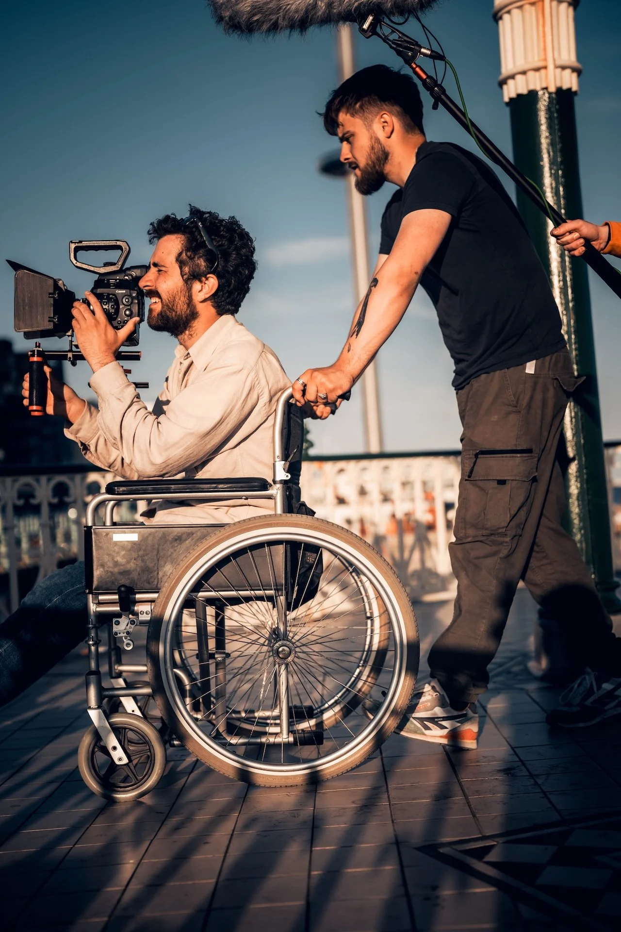 A man in a wheelchair is being pushed by another man, and they are filming a video outdoors during sunset.
